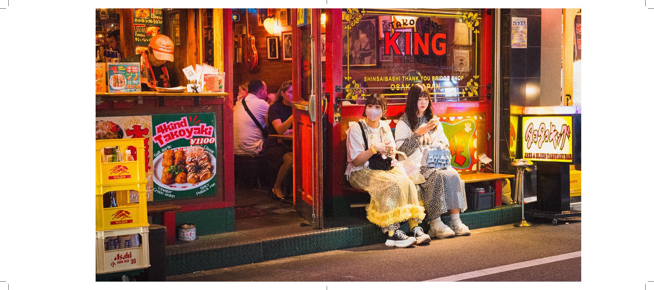 Two young women sitting outside a Japanese takoyaki restaurant at night, one wearing a face mask and the other looking at her phone, with patrons dining inside and colorful signs around.