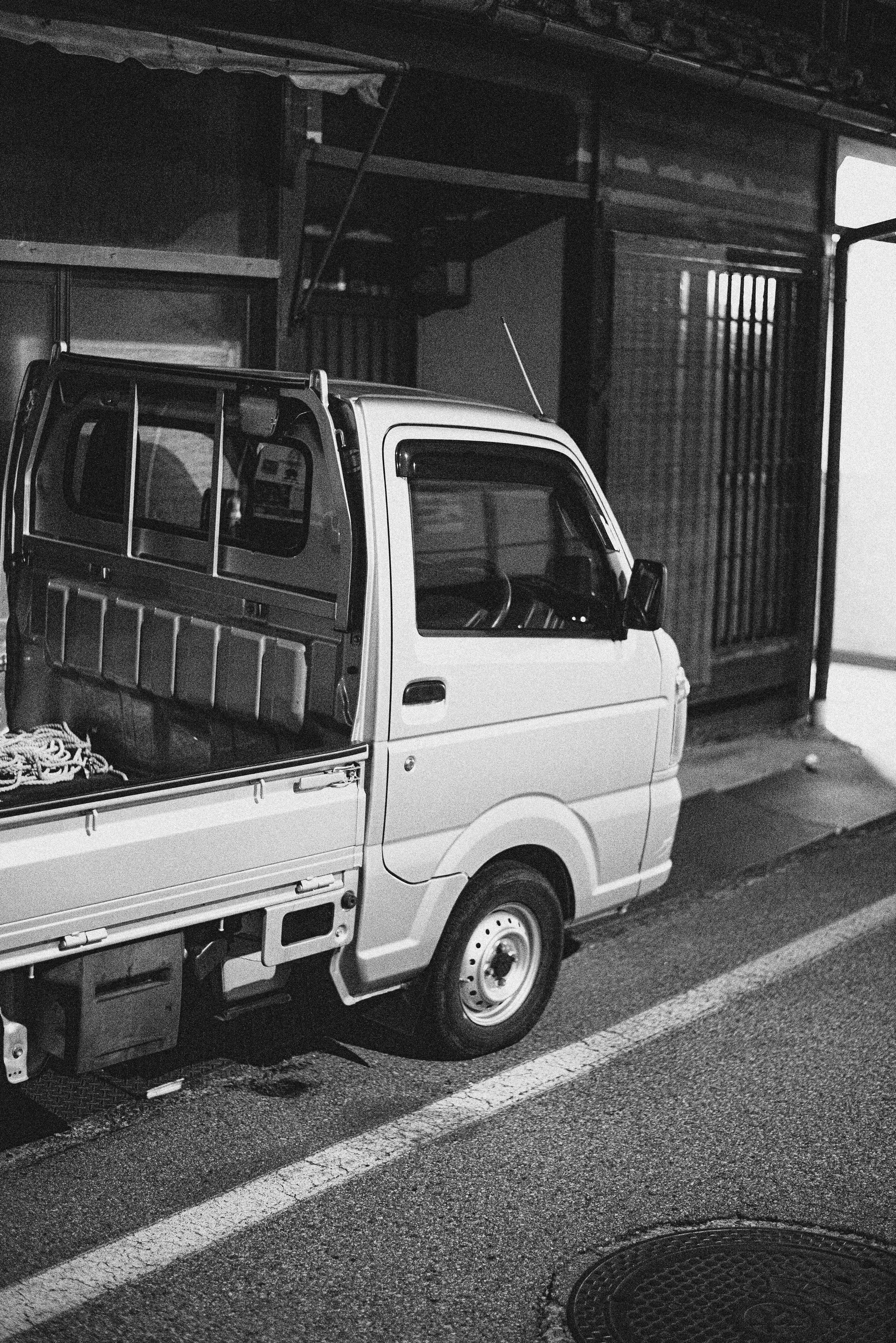 A black and white photo of a small pickup truck parked on the side of a street in front of a building or garage with sliding doors.