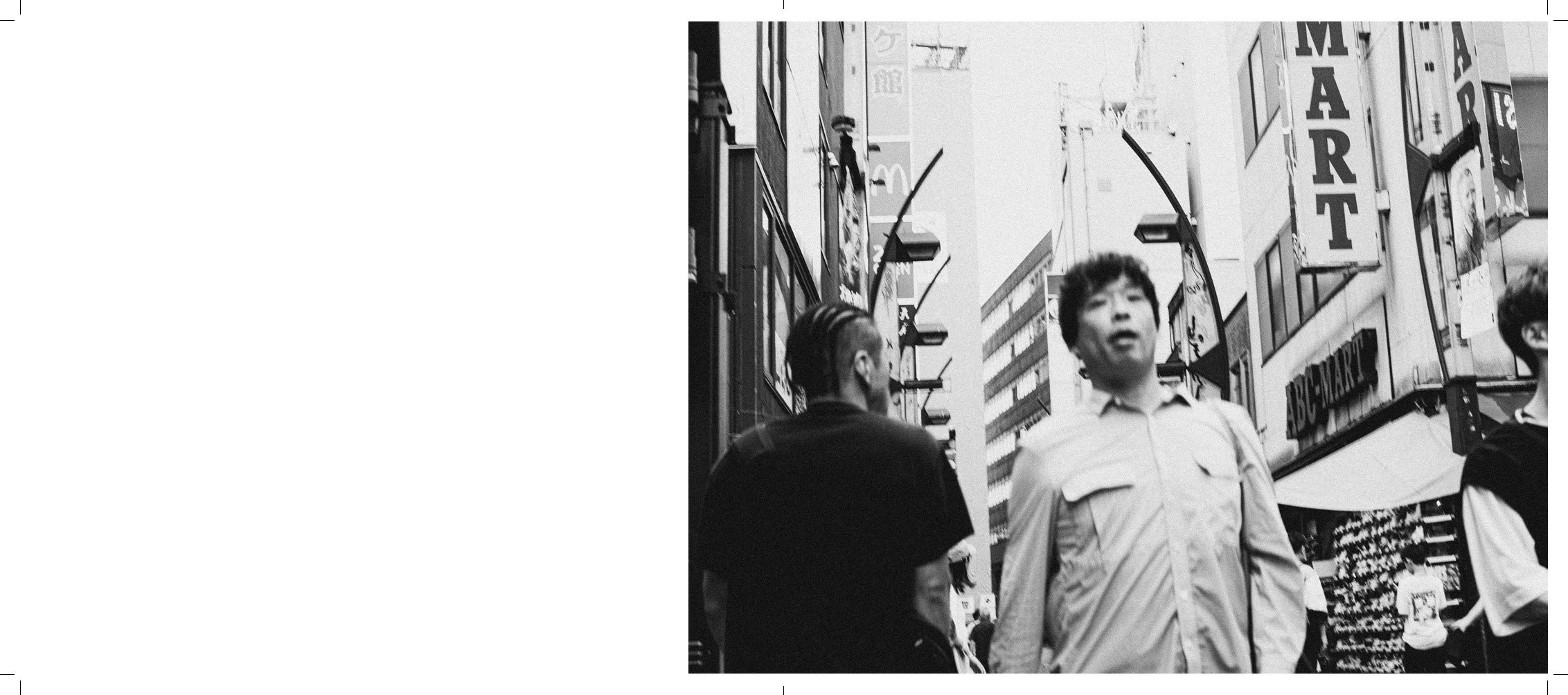 Black and white photo of people walking on a city street with tall buildings and signs, including a visible sign that says "MART". The people are engaged in conversation or looking around, with some facing the camera and others with their backs turne