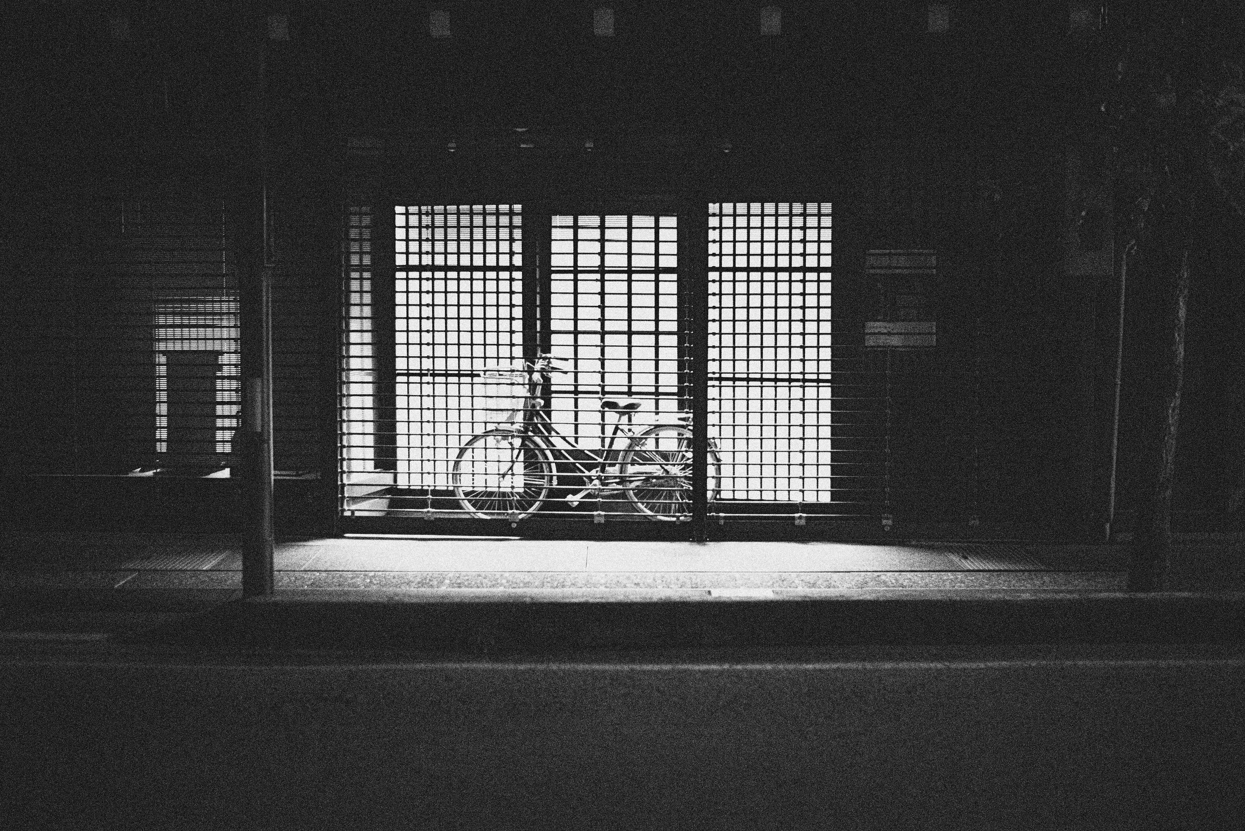 A bicycle parked behind a metal gate with a grid design, backlit by bright light coming from a window or opening, creating a silhouette effect in the dark environment.