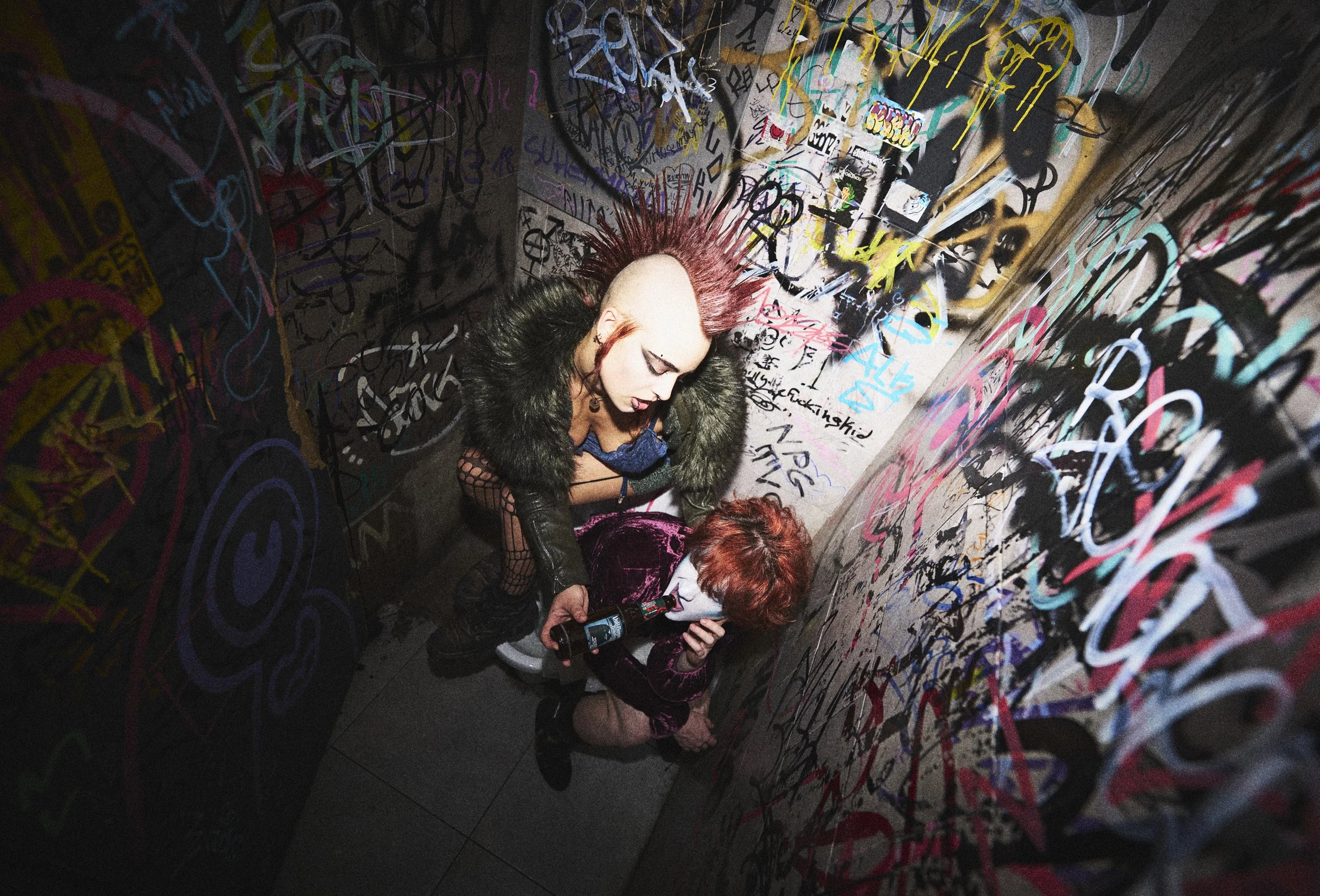 Three people in a dimly lit room with graffiti-covered walls. One person with styled hair and dark makeup is looking down at a phone, while the other two people are looking at the phone. The graffiti features various colorful scribbles and tags.