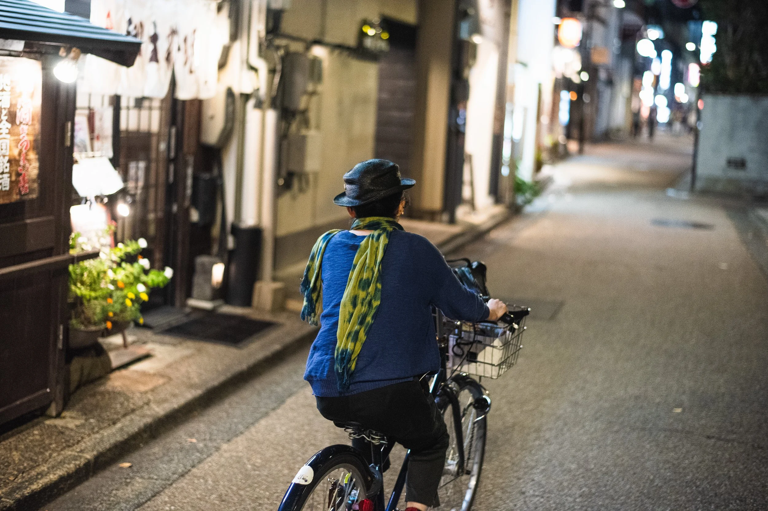 A woman riding a bicycle on a quiet, dimly lit street at night, wearing a black hat, a blue jacket, and a green scarf.