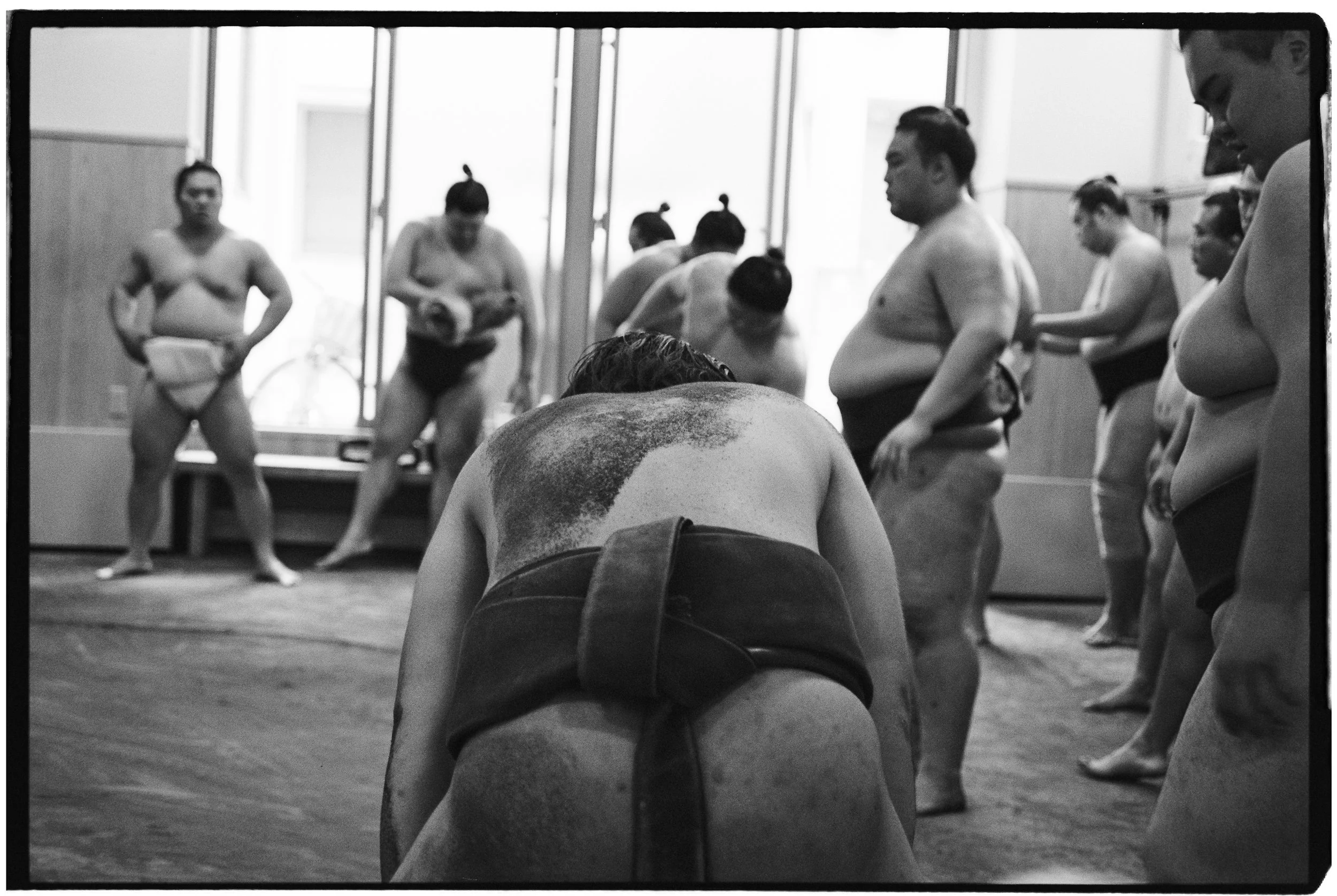 Sumo wrestlers practicing in a dojo, with a wrestler bowing in the foreground and others standing or preparing in the background.