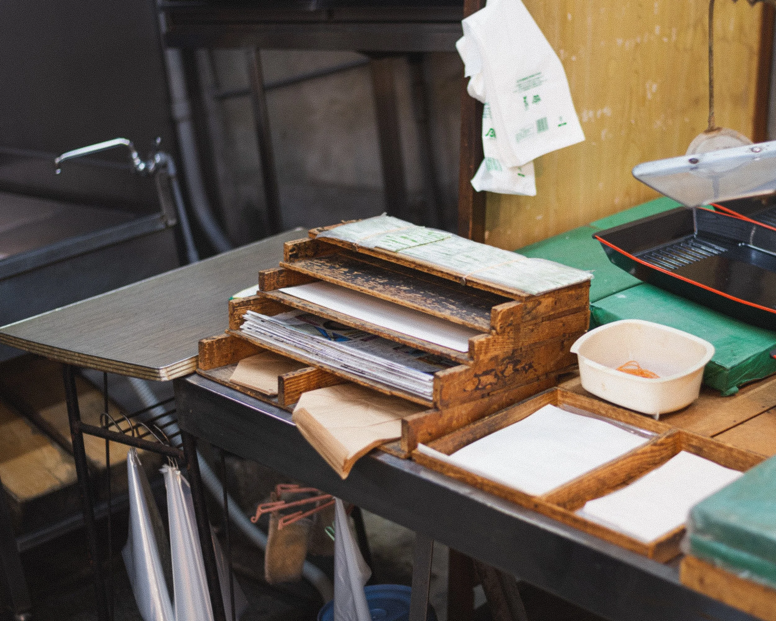 A wooden desk with stacked old wooden trays filled with papers, a small white plastic container, and a black gridded case. Items hanging below the desk, a green mat on the right, and various objects on the background including a dark countertop with 