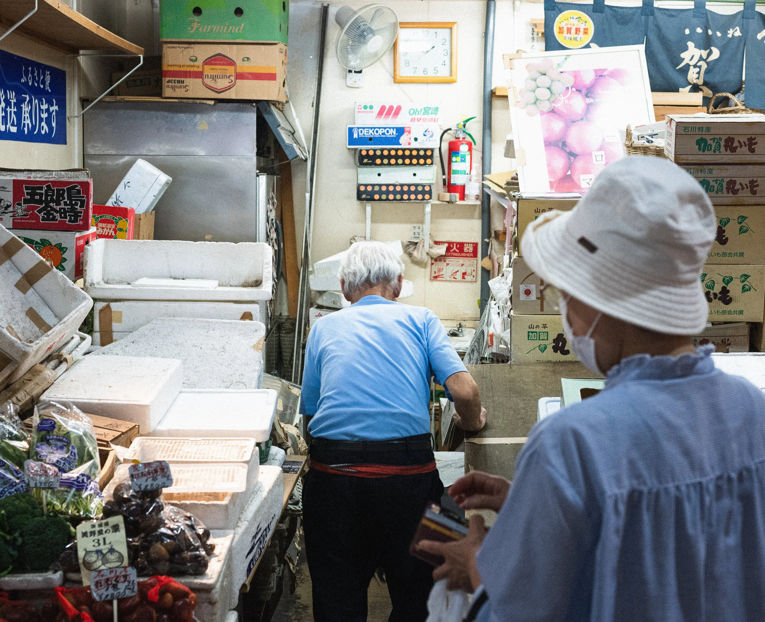 Two elderly women in face masks shopping at a small market stall with various fruits and vegetables, with signs and boxes in the background.