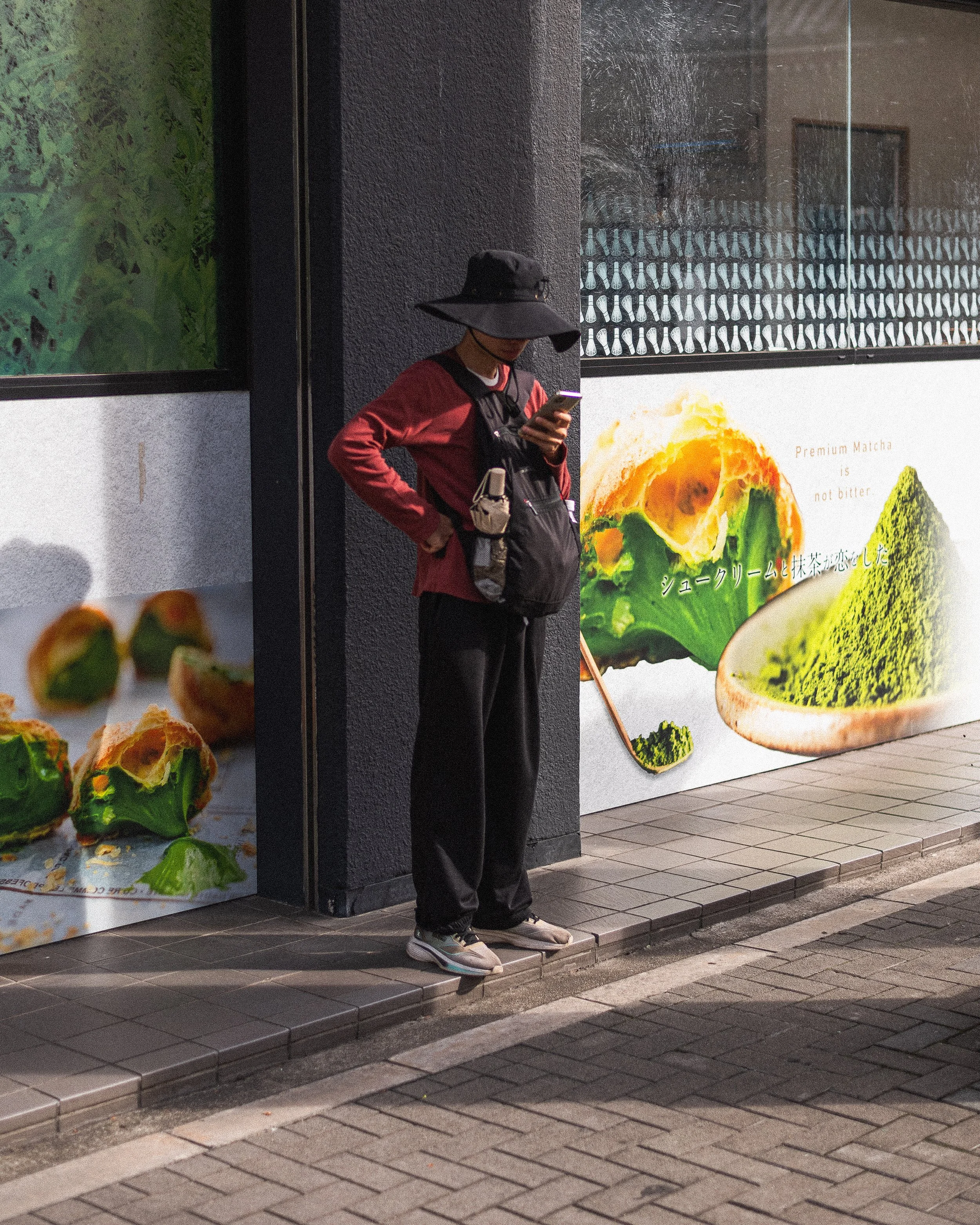 A person standing on a city sidewalk at night, wearing a wide-brimmed black hat, red long-sleeve shirt, black pants, and sneakers, looking at a smartphone. They have a backpack with small pouches on their chest and waist.