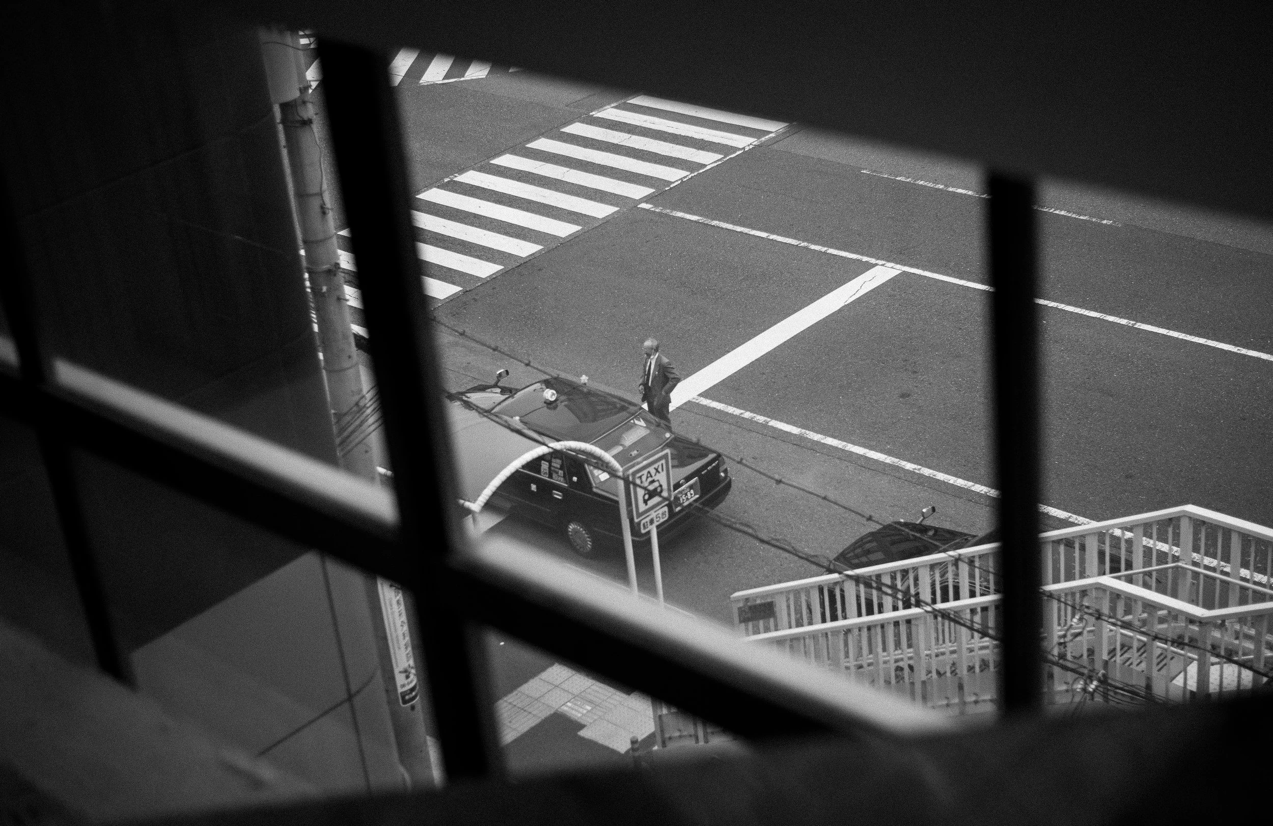 Black and white photo taken from an elevated vantage point showing a man in a suit and fedora hat walking near a taxi on a city street, with street markings including crosswalks and lanes visible.