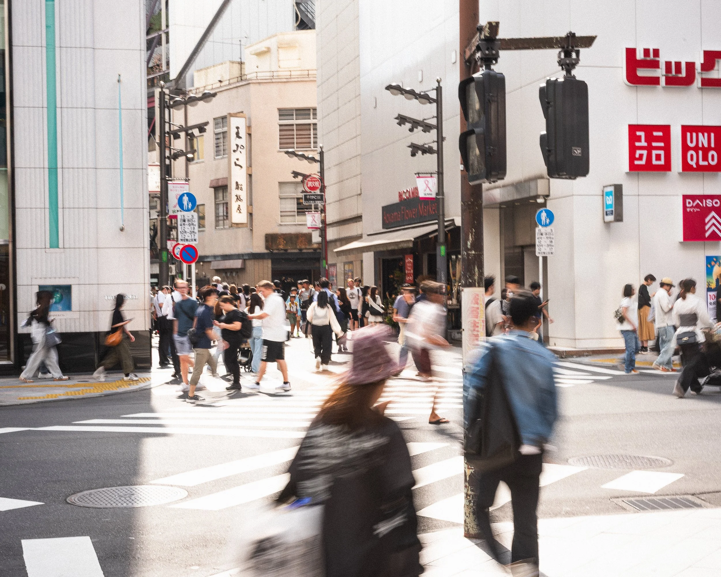 Urban street scene with many pedestrians crossing a crosswalk, surrounded by commercial buildings and signs, including UNIQLO, DAISO, and others.