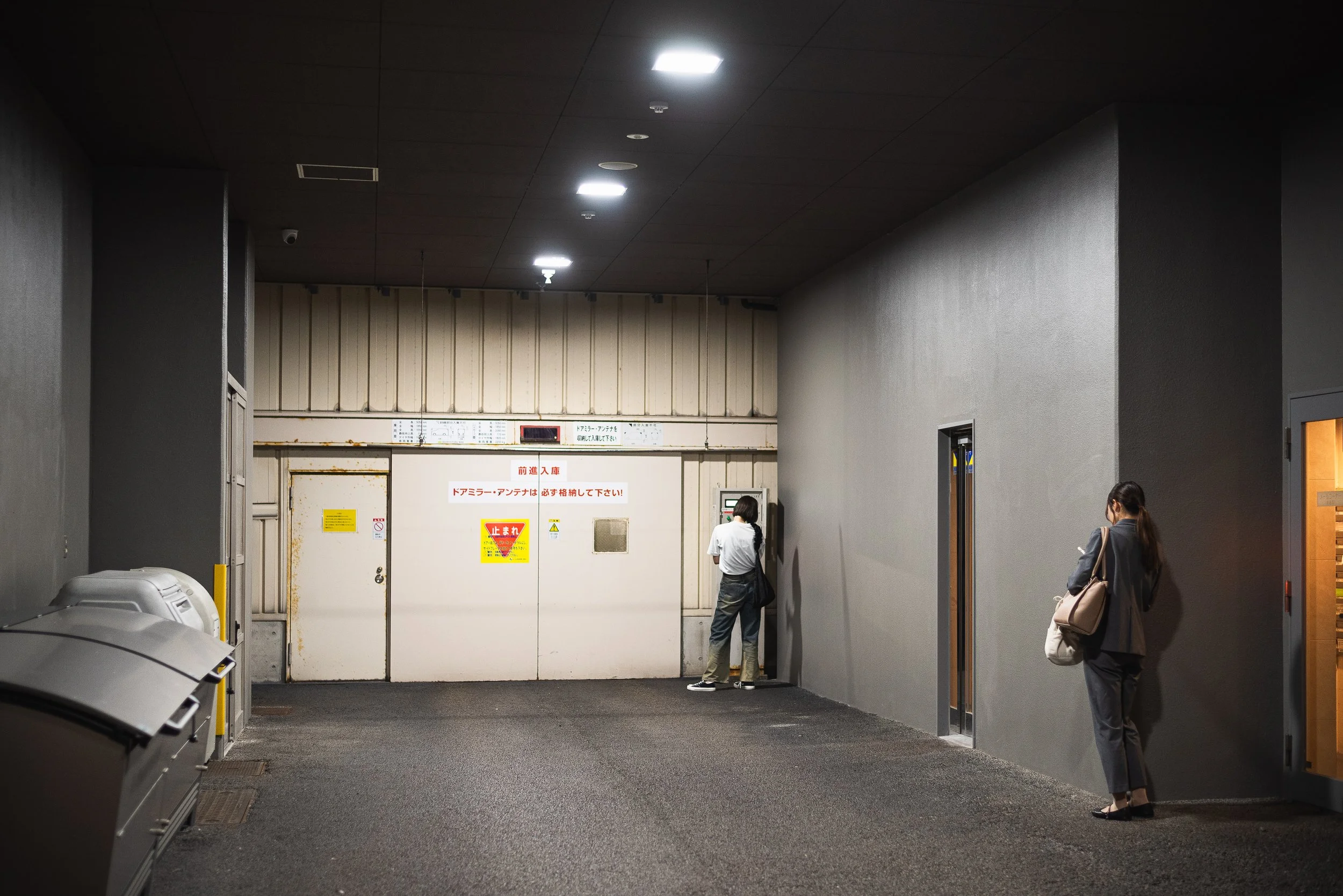 Indoor scene with two women, one using a payphone near a door with warning signs, the other standing against a gray wall, illuminated ceiling lights, and a brown door on the right.