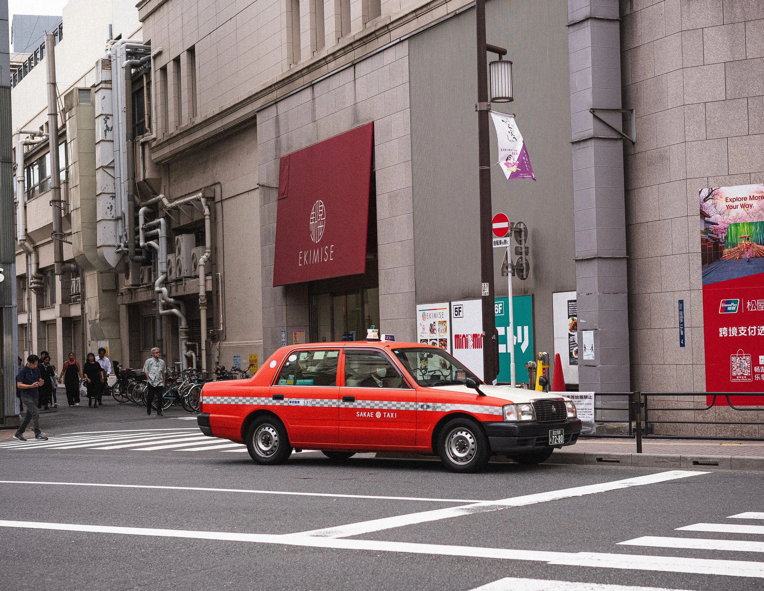 A red and white taxi cab parked on the side of the street in front of a building with large signs and several pedestrians walking, some bicycles parked nearby, in an urban setting.