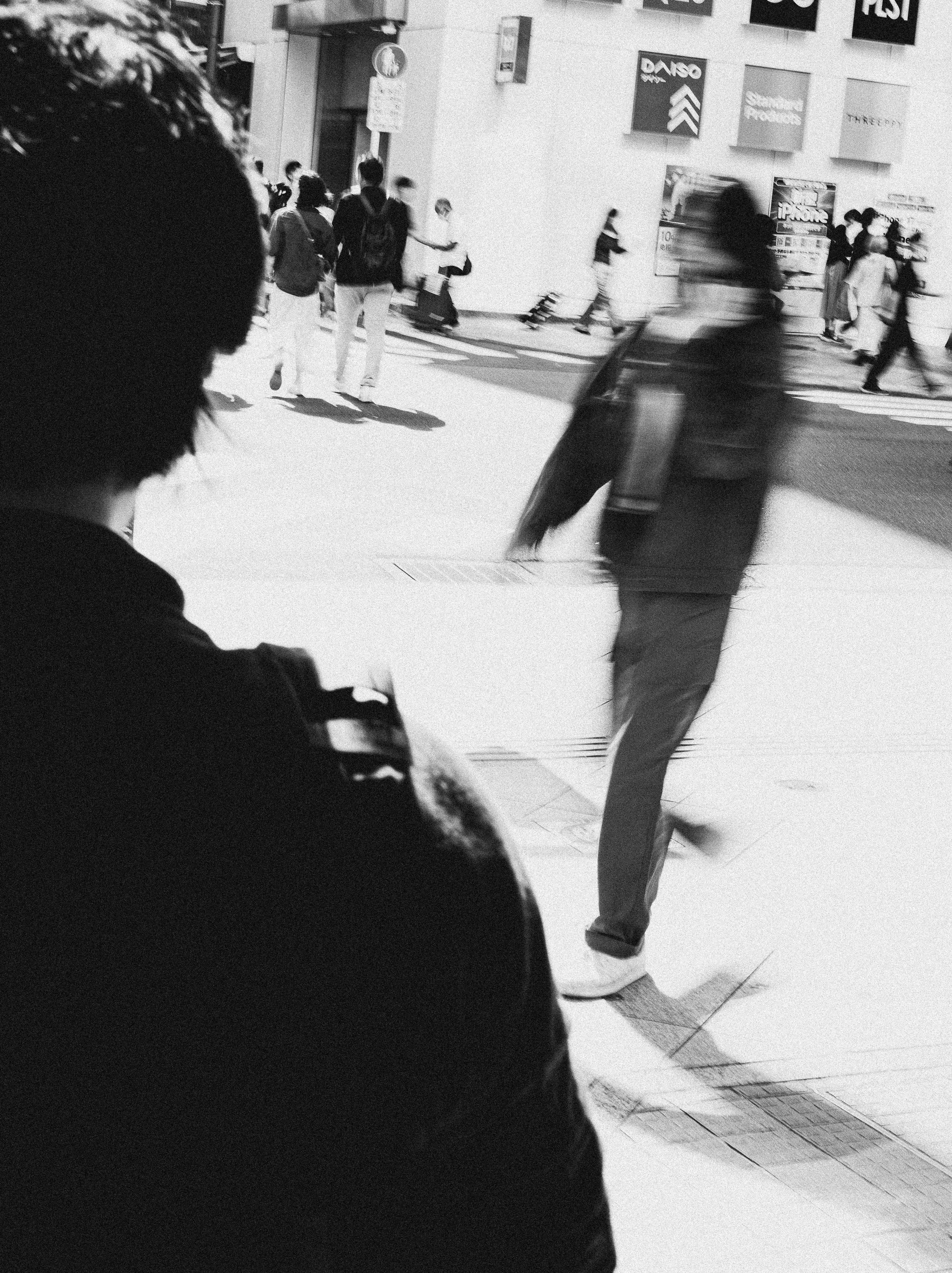 Black and white photo of a busy city street with people walking. In the foreground, a person with short hair looks at their phone, while a man with a skateboard passes by, slightly blurry.