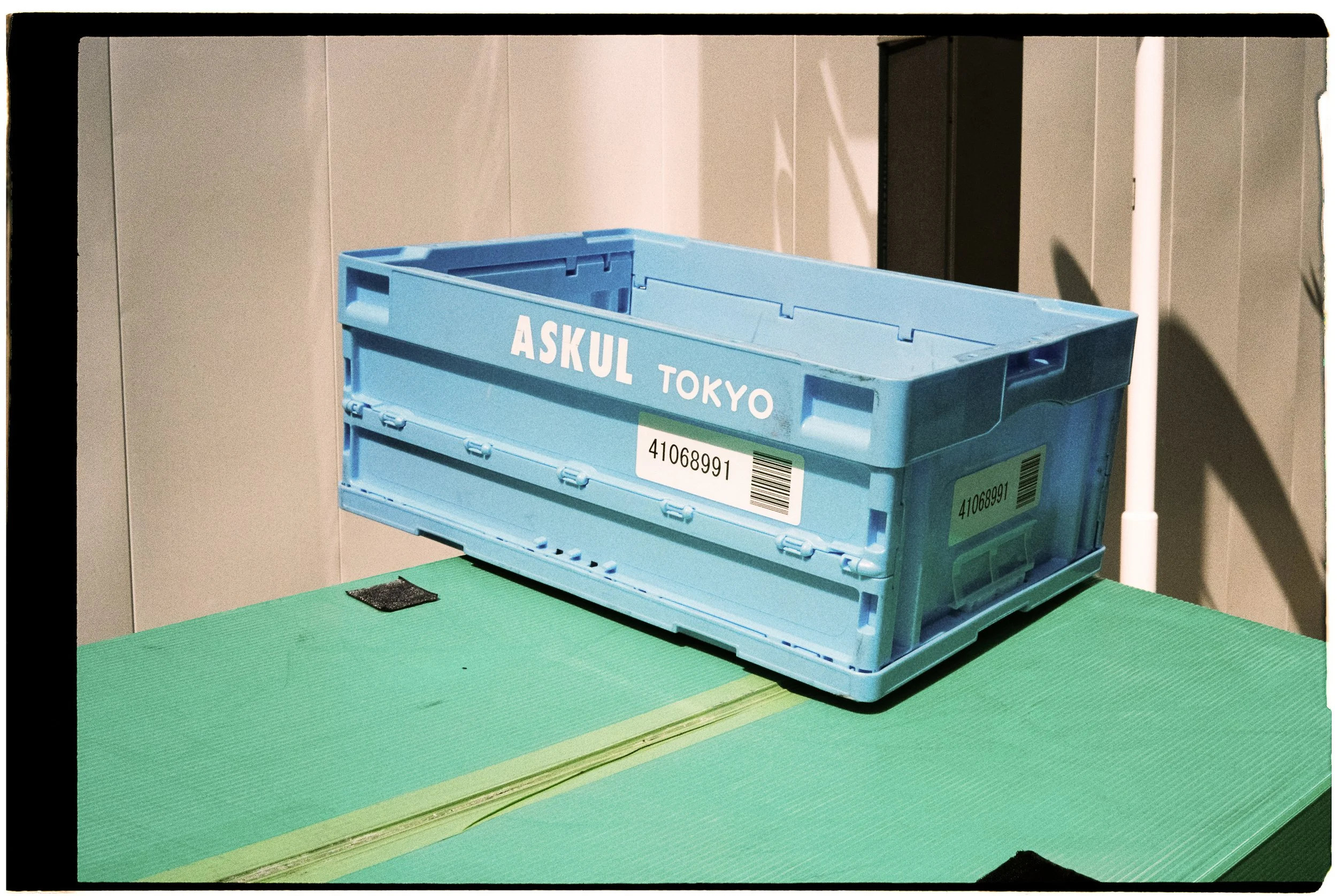 A blue plastic crate labeled with 'ASKUL TOKYO' and barcodes, placed on a green table surface against a light-colored wall.