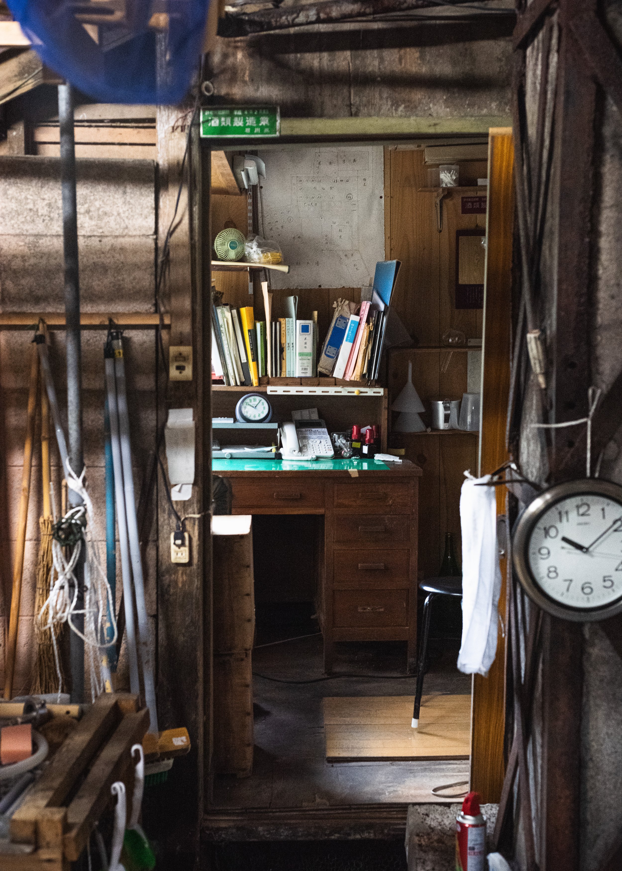 A cluttered home office seen through a mirror frame, featuring a wooden desk with various items including a clock, phones, and office supplies, surrounded by bookshelves and personal belongings.