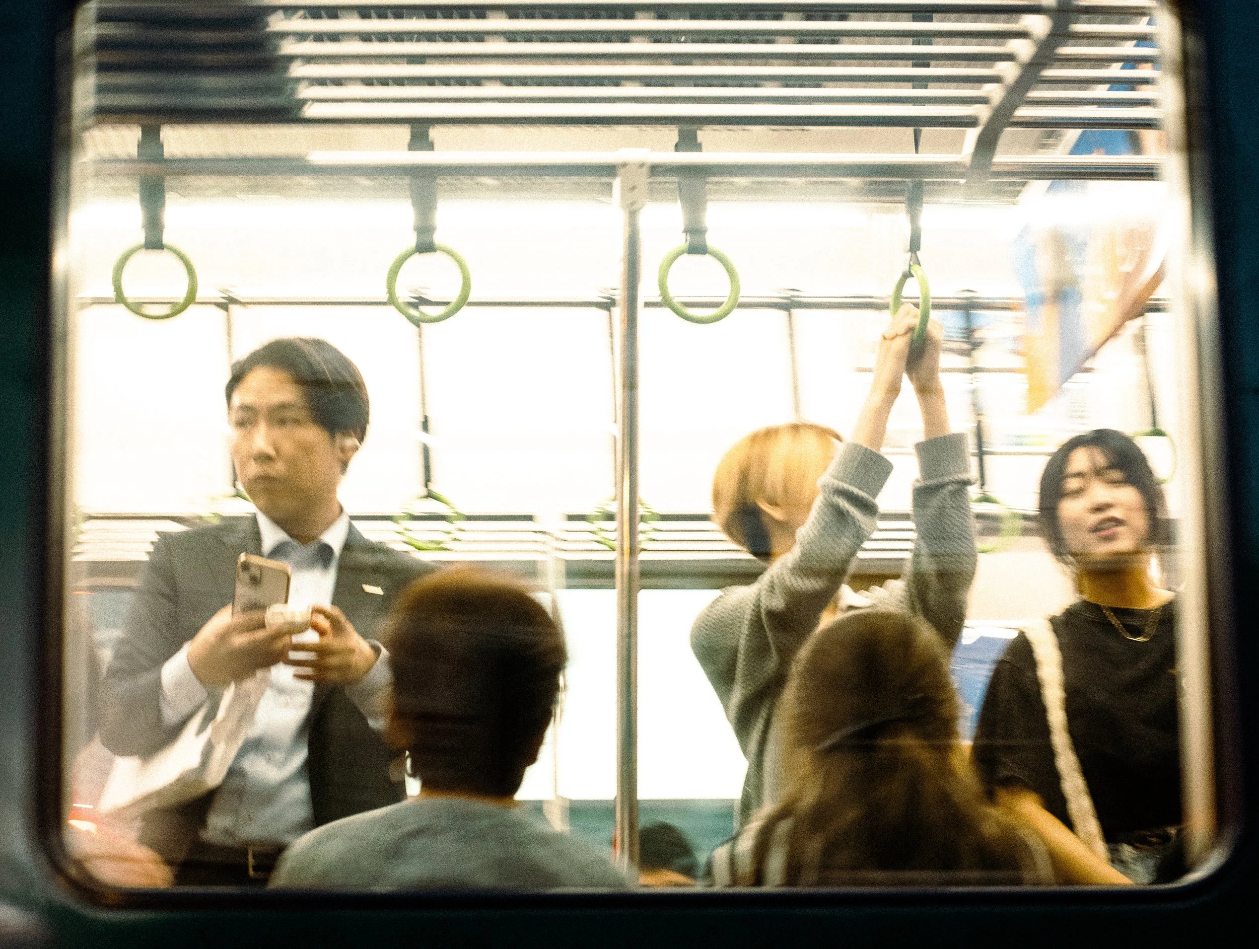 People standing and sitting inside a subway train, some holding onto handrails and one person using a smartphone, viewed through a train window.