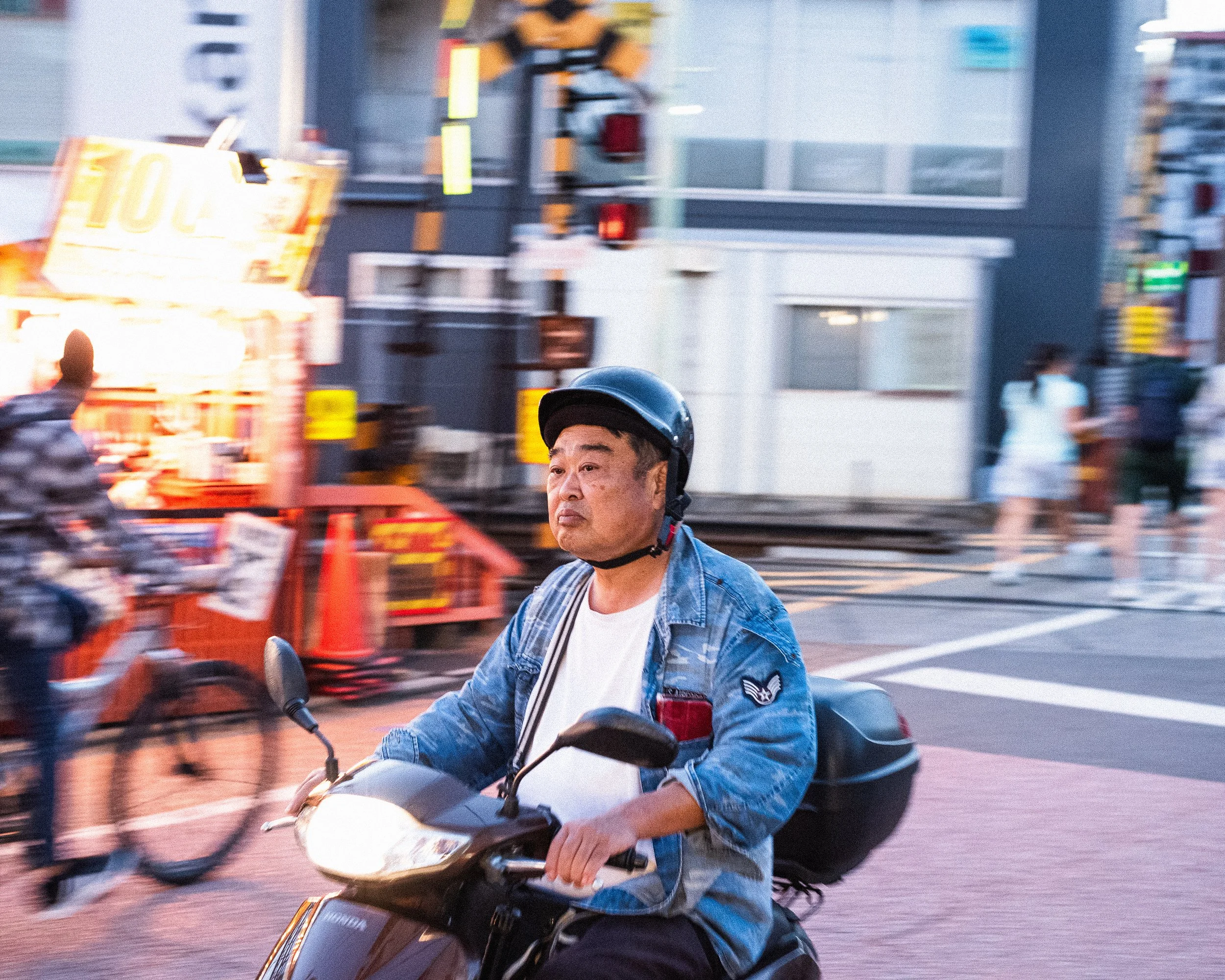 A man riding a motorcycle on a city street. The man is wearing a helmet and a denim jacket, and other pedestrians and cyclists are visible in the background.