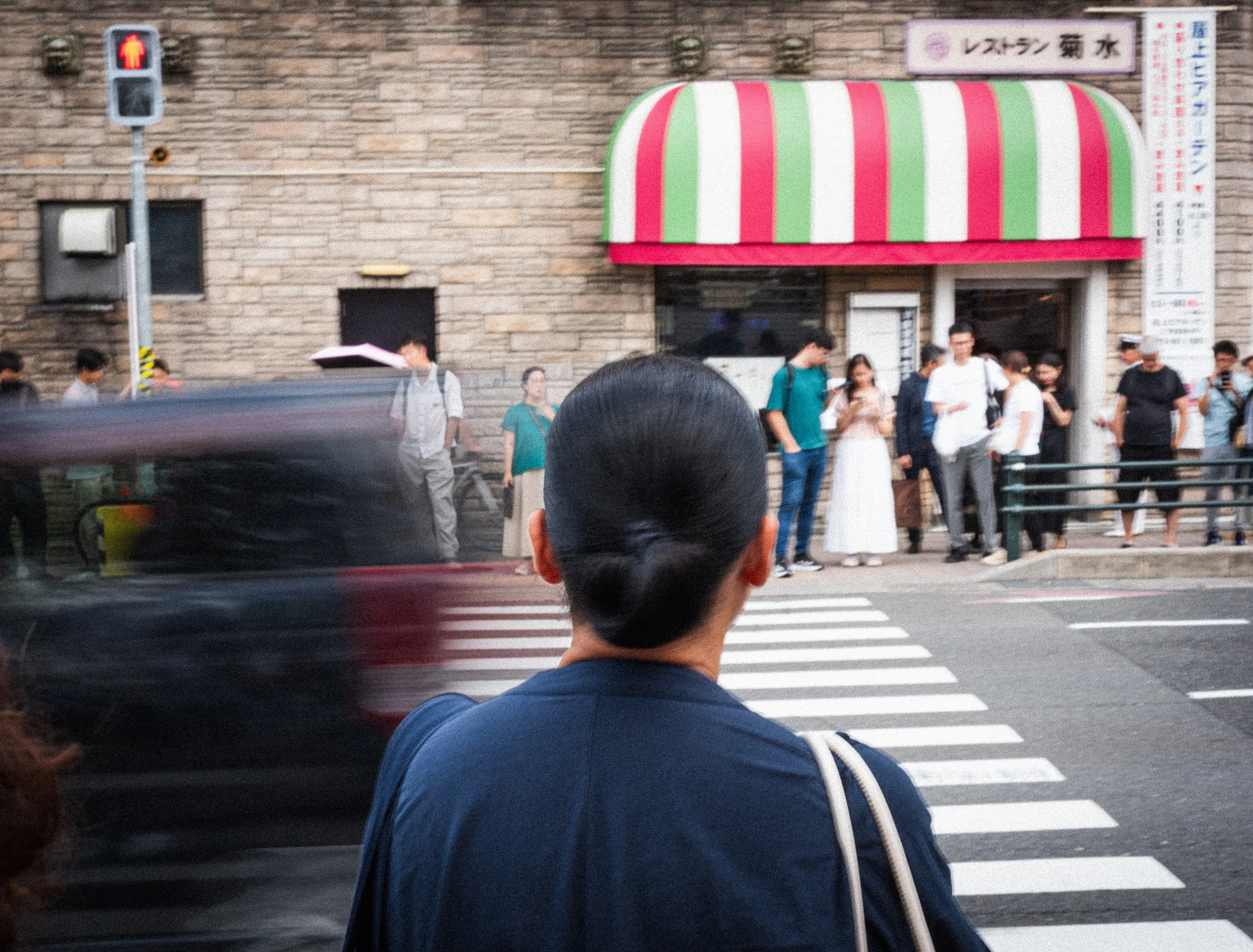 A woman with dark hair in a bun is standing at a crosswalk, watching a black car pass by. In the background, there is a group of people standing outside a building with a red and green striped awning and a sign with Japanese characters. A pedestrian 