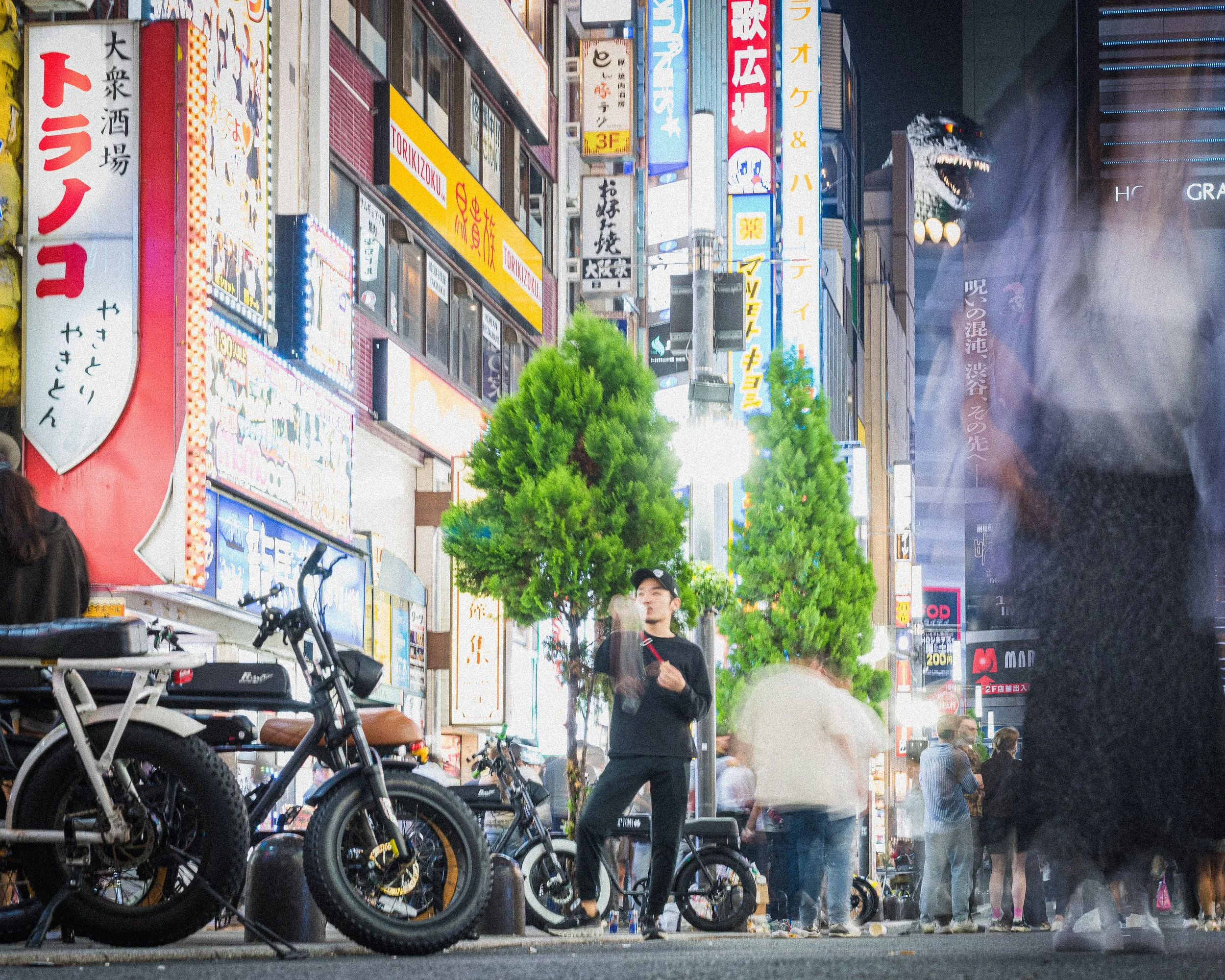 A lively city street scene at night in Japan, with brightly lit signs in Japanese, bicycles parked along the sidewalk, a person standing and smoking, and a dragon head decoration projecting from a building in the background.