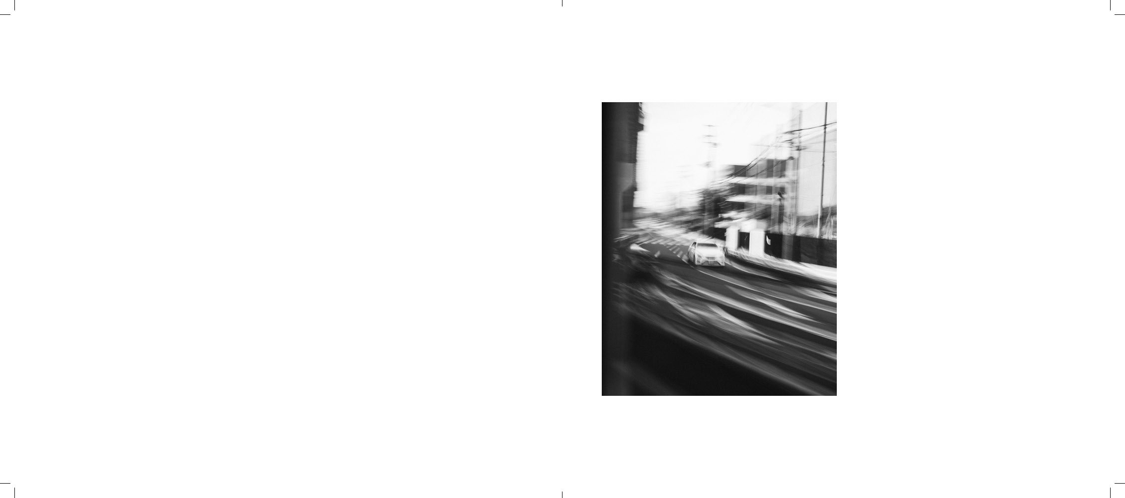 Black and white photo of a city street seen from a moving vehicle, with buildings, power lines, and a white car in the distance.