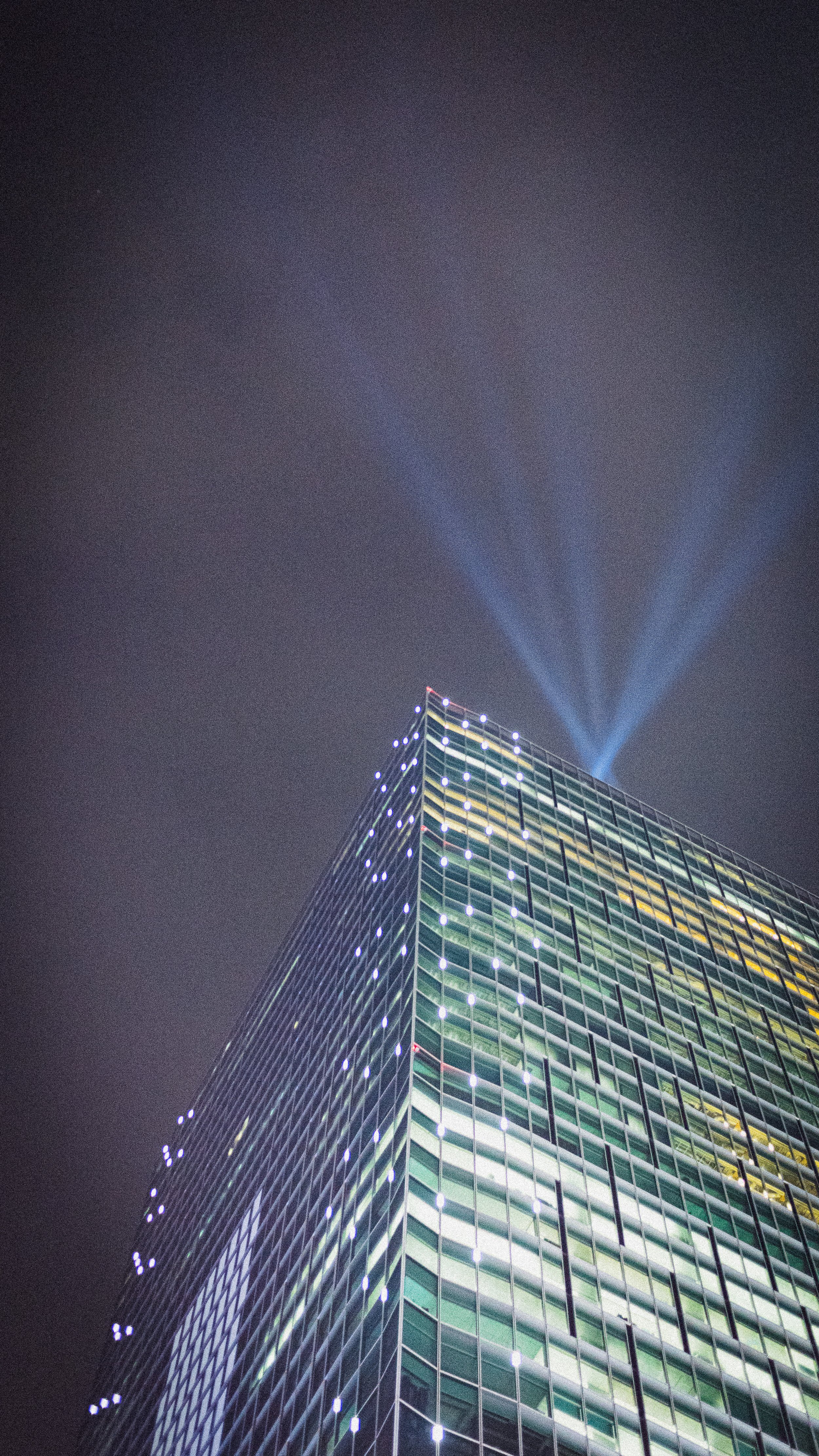 Nighttime view of a tall modern glass building with lights in the windows and blue spotlights shining from the top into the dark sky.