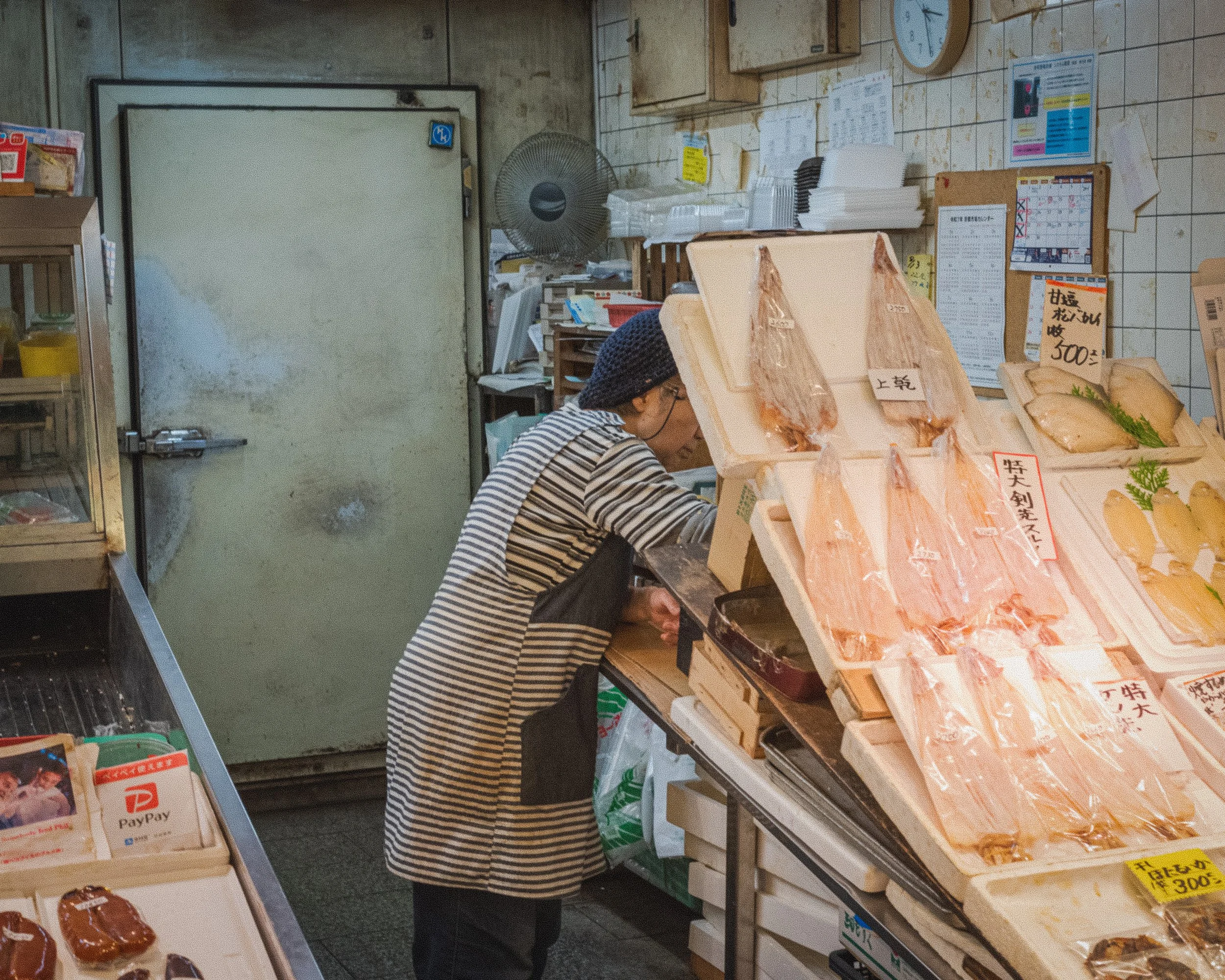 A woman in a striped shirt and a blue head covering shops for fish at a market stall with dried fish displayed on foam trays. The shop has tiled walls, a clock, and various papers and signs pinned up.