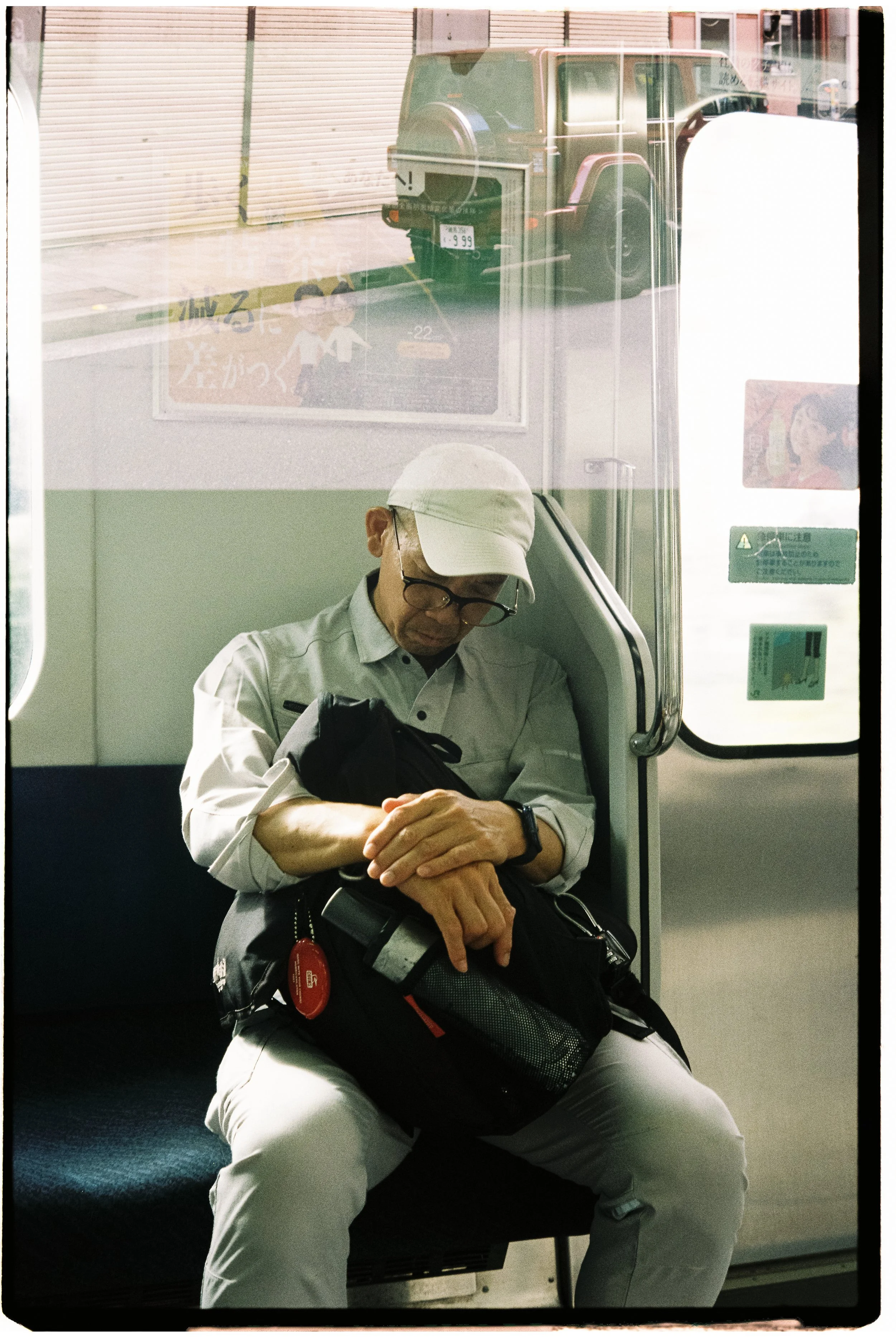 A man sitting on a train, resting with his eyes closed, wearing a white cap, glasses, and light-colored clothing, holding a backpack on his lap.