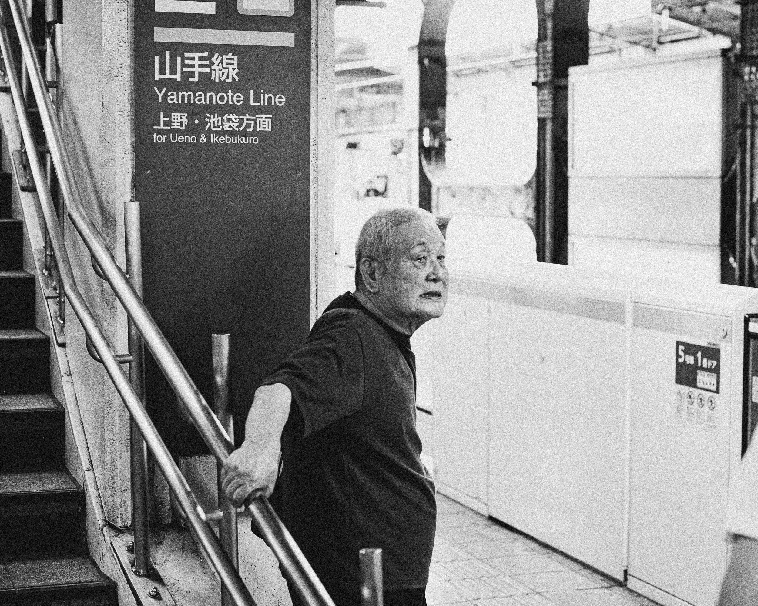 Black and white photo of an elderly man with gray hair, wearing a dark shirt, standing next to a stairway on the left. He is holding the handrail and looking to his right, with a sign for the Yamanote Line in Japanese and English visible behind him. 