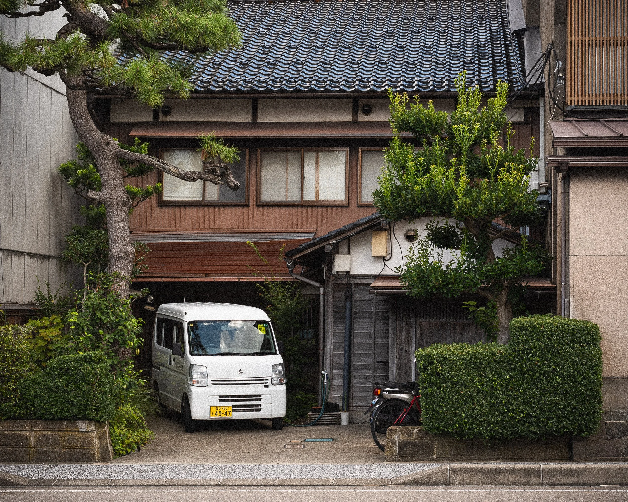 A quiet residential scene featuring a traditional Japanese house with a tiled roof, surrounded by lush greenery including trees and bushes. A white Suzuki small van is parked in the driveway, with a black bicycle beside it. The house has a mix of woo