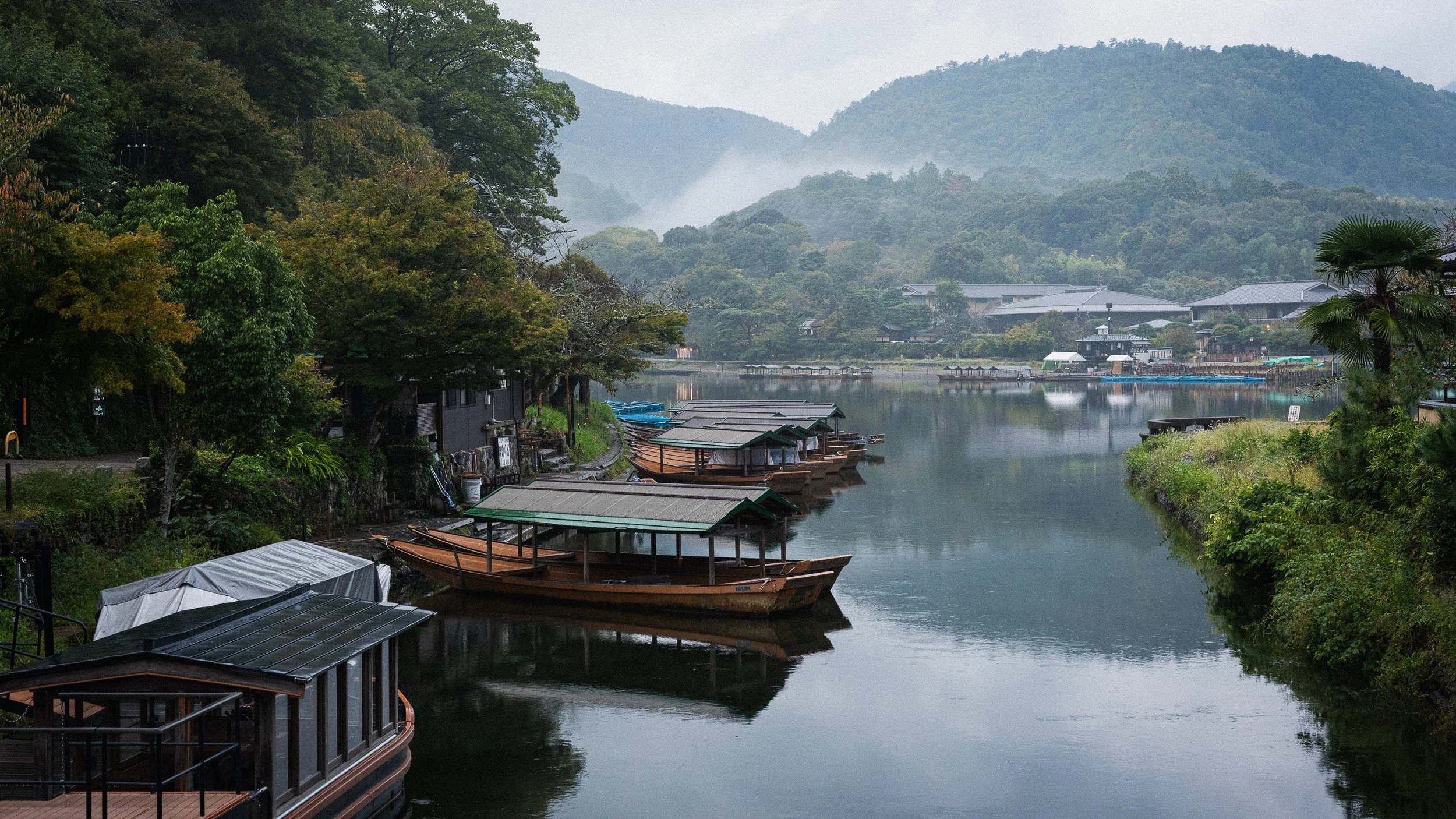 A tranquil river scene with traditional wooden boats with green-roofed shelters docked along the riverbank. The surrounding area is lush with trees and mountain hills in the background, with mist over the mountains and a cloudy sky.