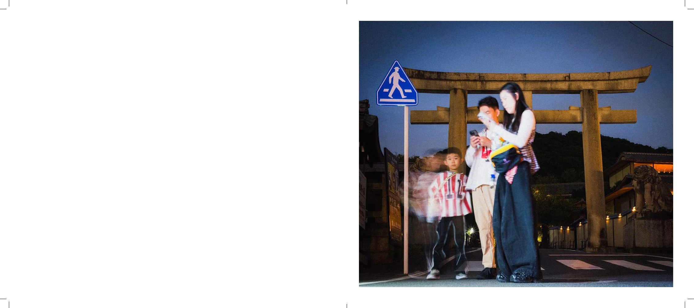 People crossing the street at night in front of a traditional Japanese torii gate with a blue pedestrian crossing sign nearby.