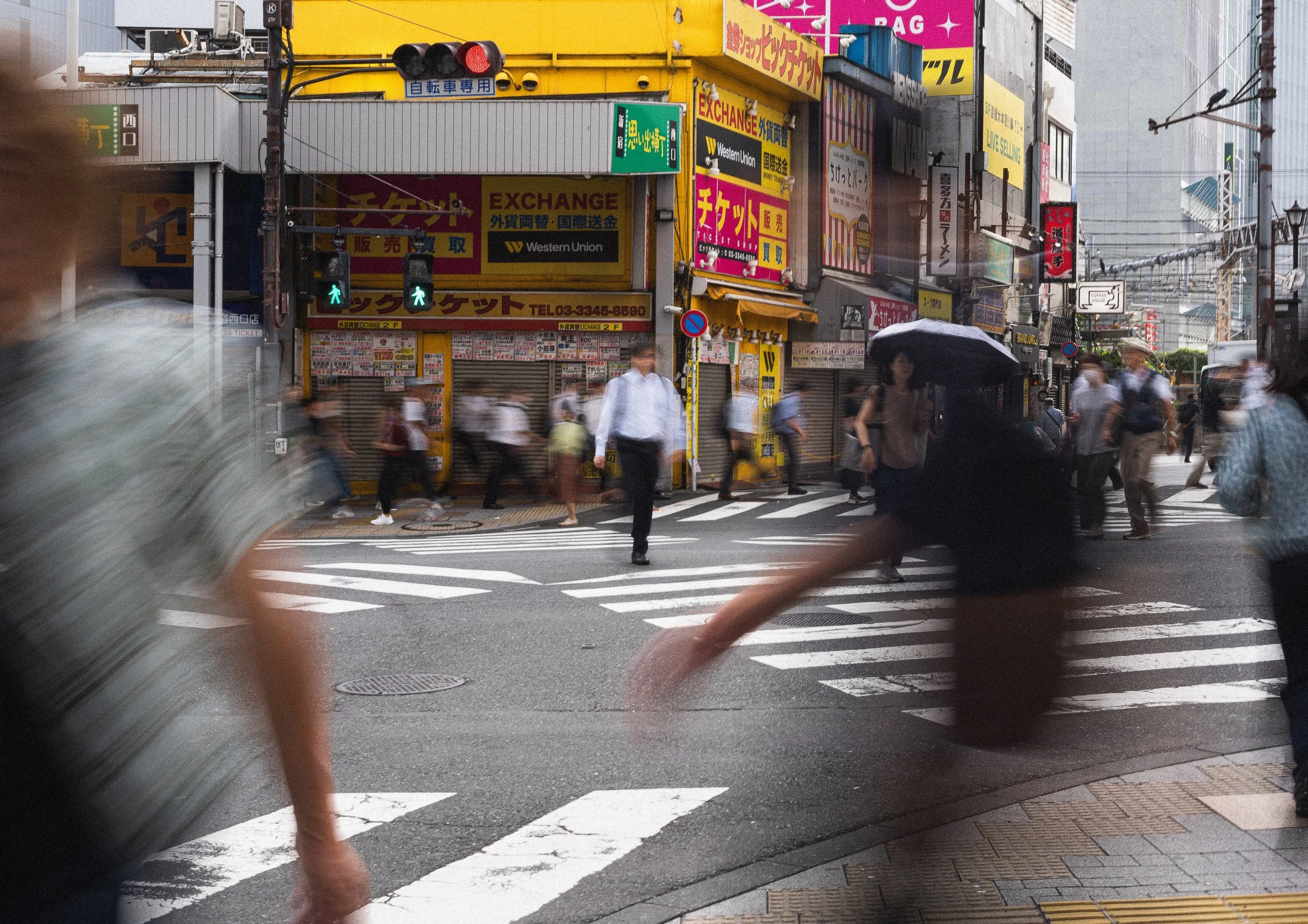 A busy city crosswalk on a rainy day with blurred pedestrians rushing across the street. People hold umbrellas, and storefronts with colorful signs are visible in the background.
