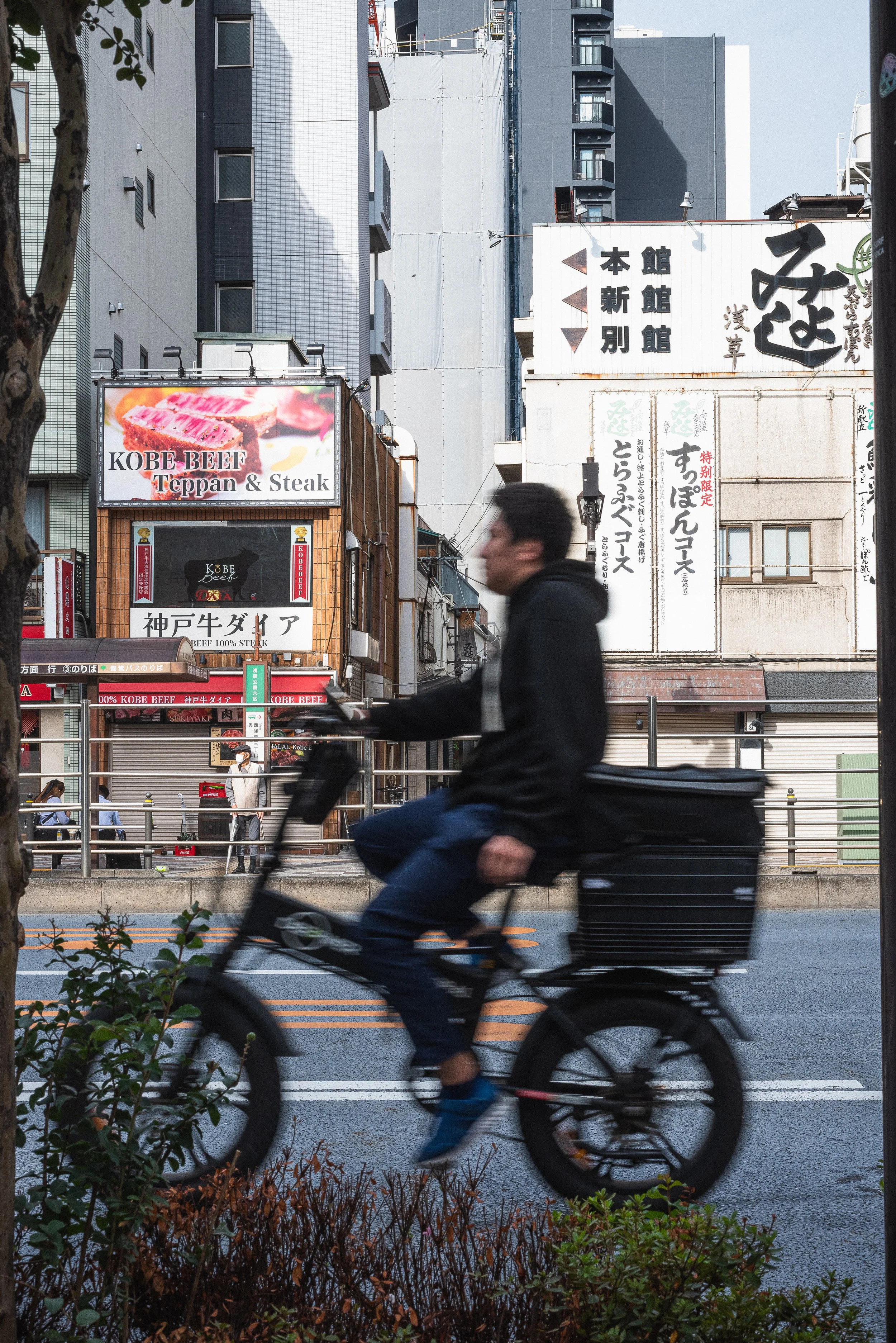 A person riding a bicycle on a city street with buildings and signs in Japanese and English in the background.