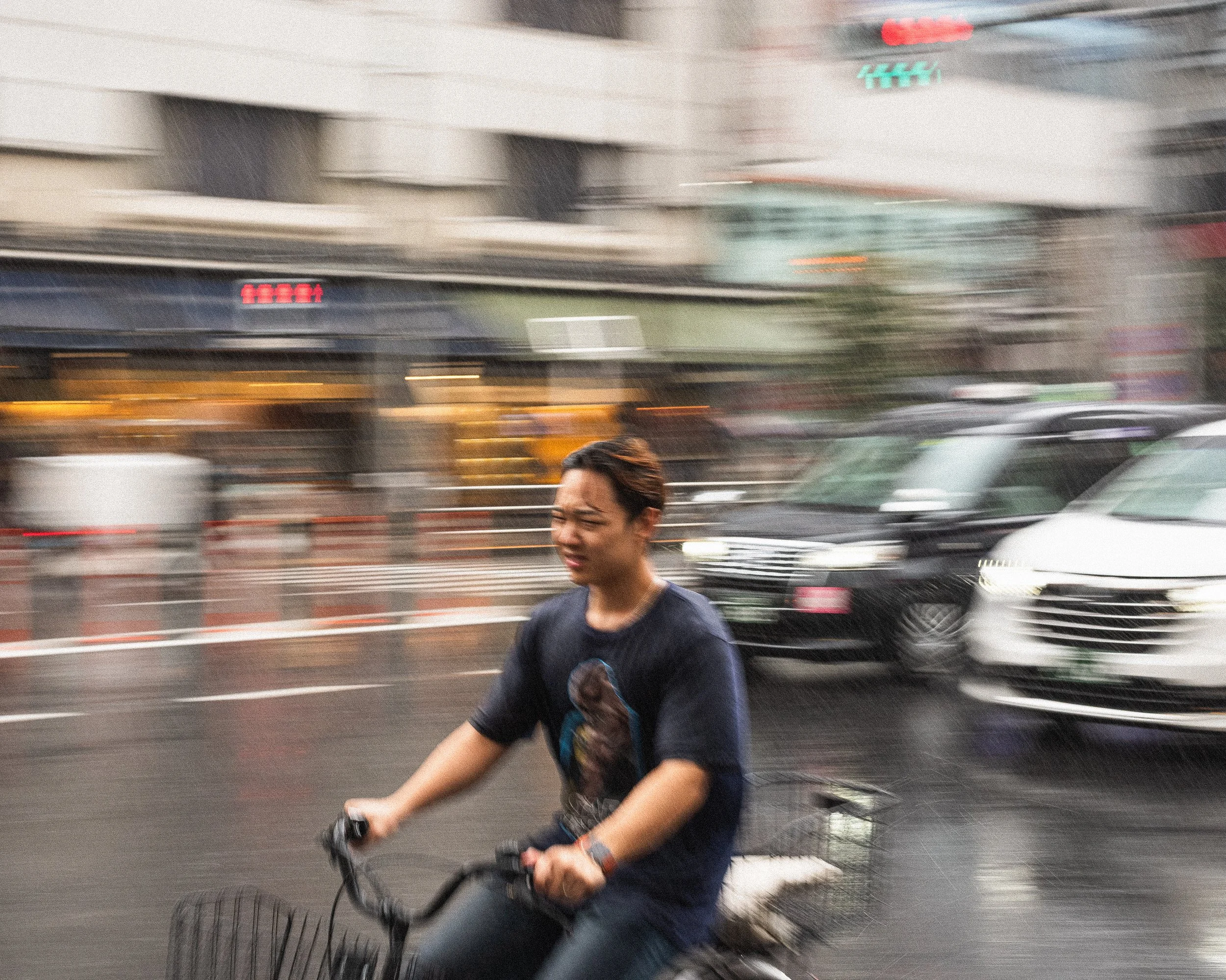 A person riding a bicycle on a city street during rain, with blurred cars and buildings in the background.