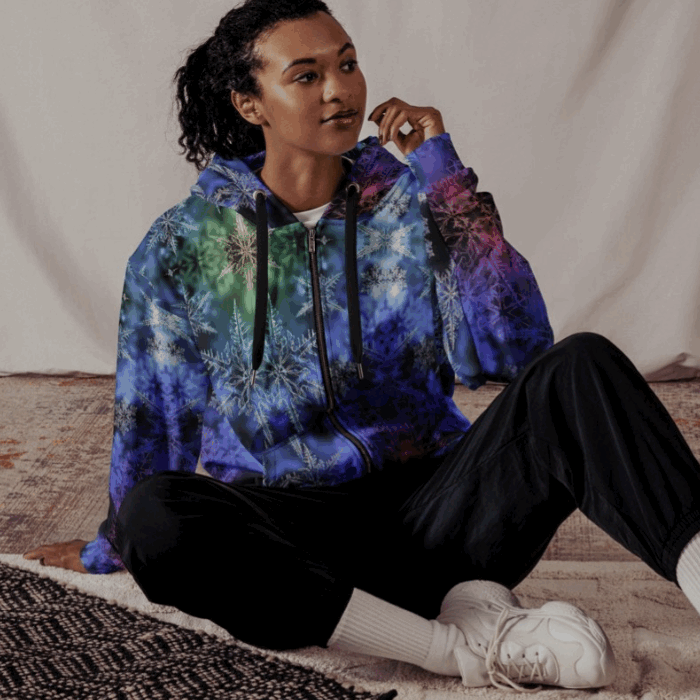 Young woman sitting on a beige carpet, wearing a colorful galaxy-themed hoodie, black pants, white sneakers, and white socks, with a neutral background.