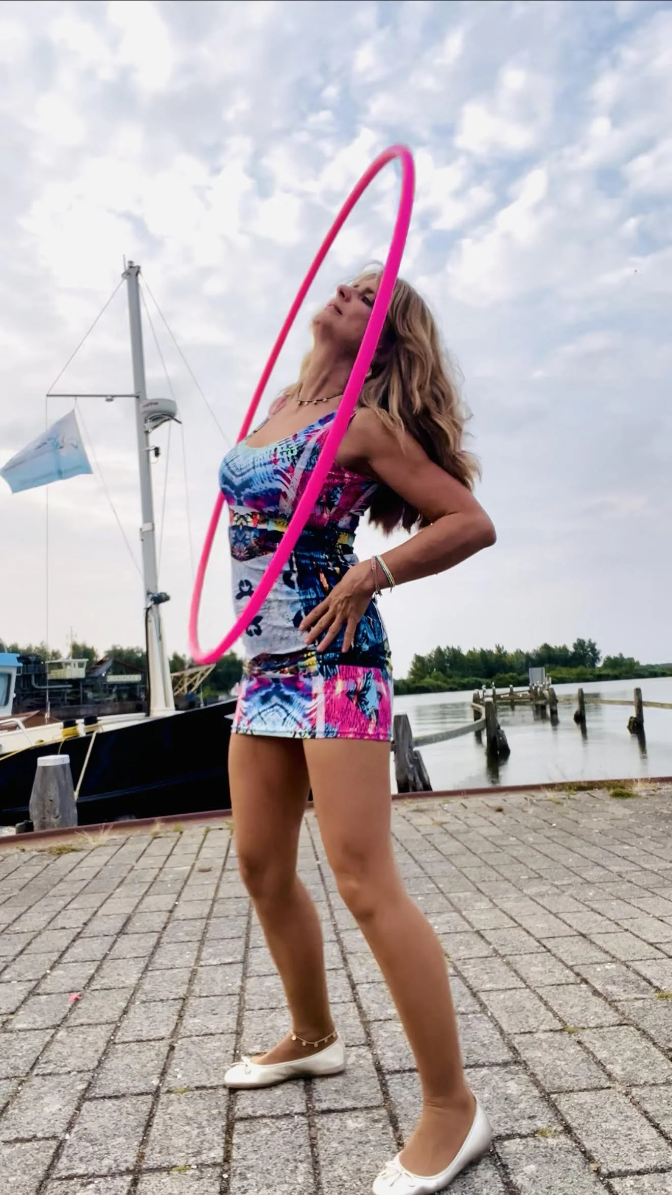 Hulamyhoop outdoors near a marina, dressed in Nigerian Whirleytimes dress, playing ,boob hooping with a pink hula hoop.