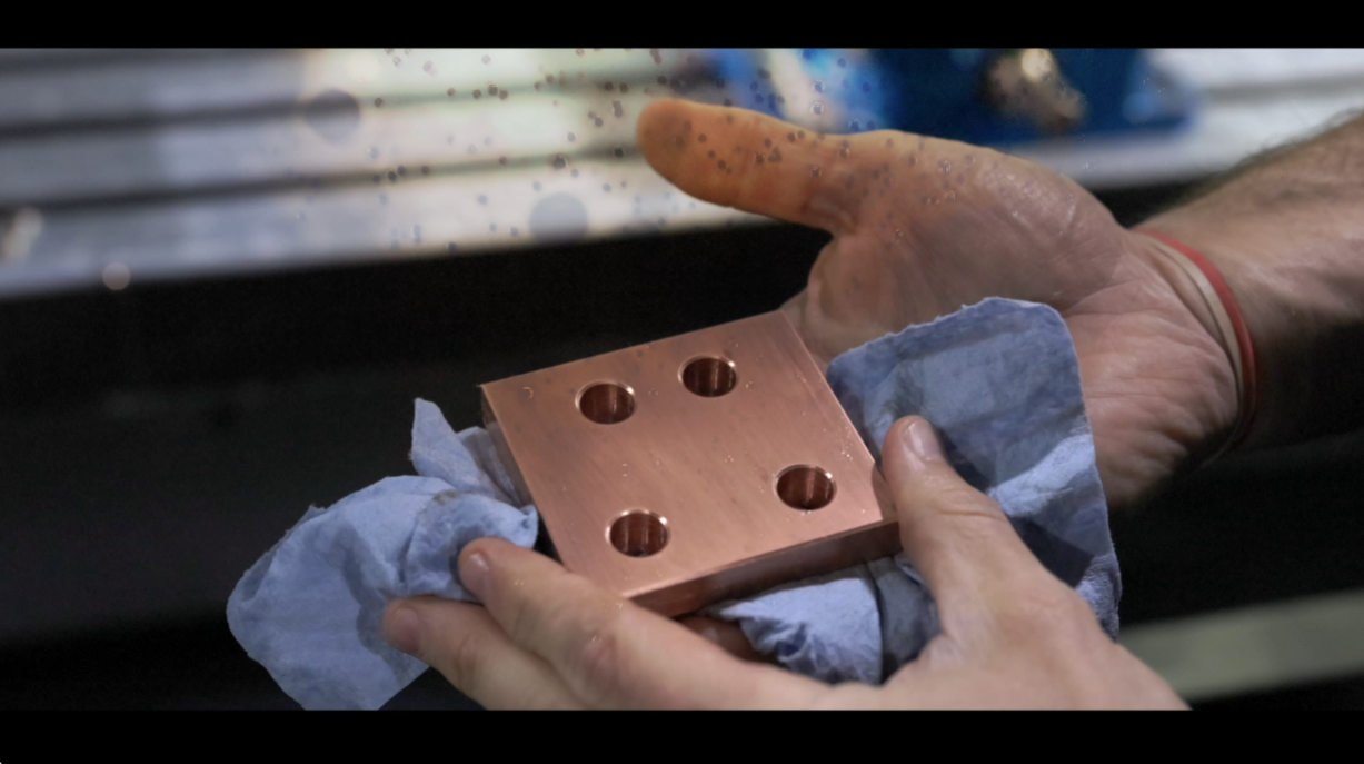 Person holding a polished copper block with four holes, using a blue cloth, industrial setting.