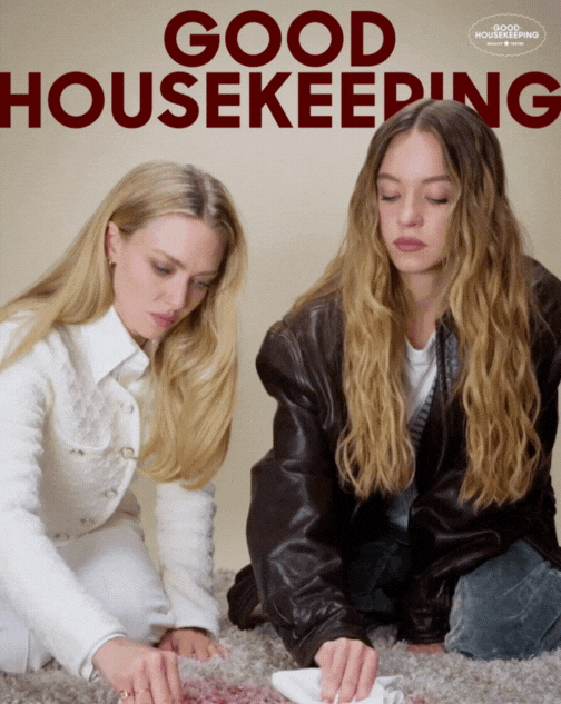 Two young women sitting on the carpet, engaging in house cleaning with a cloth and cleaning supplies, in front of a wall with the words 'Good Housekeeping' in large red letters.