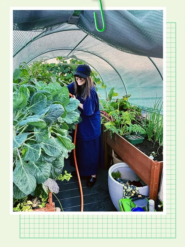 A woman smiling inside a small greenhouse or hoop house, watering plants. She is wearing a navy blue cap and glasses, surrounded by various leafy vegetables and herbs, with gardening tools nearby.