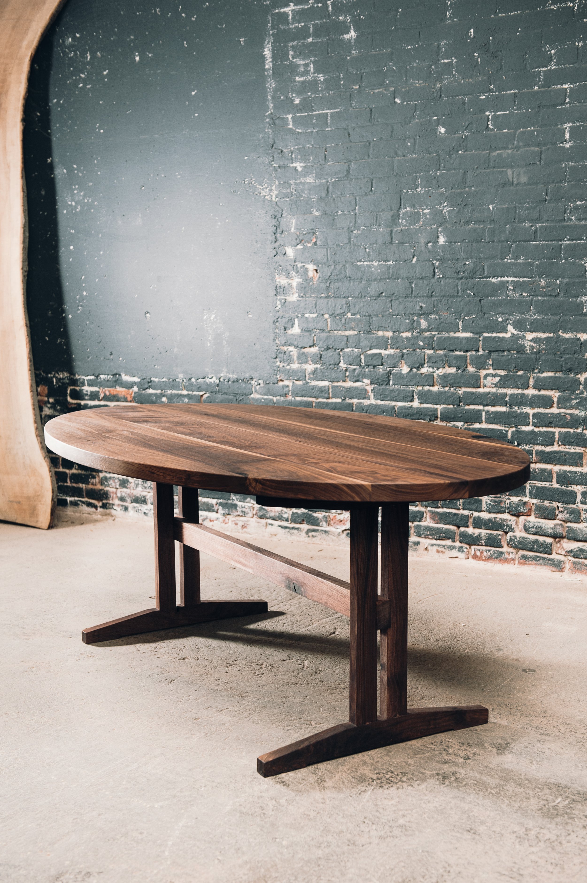A wooden dining table with a curved oval top and sturdy base, set against a dark brick wall.