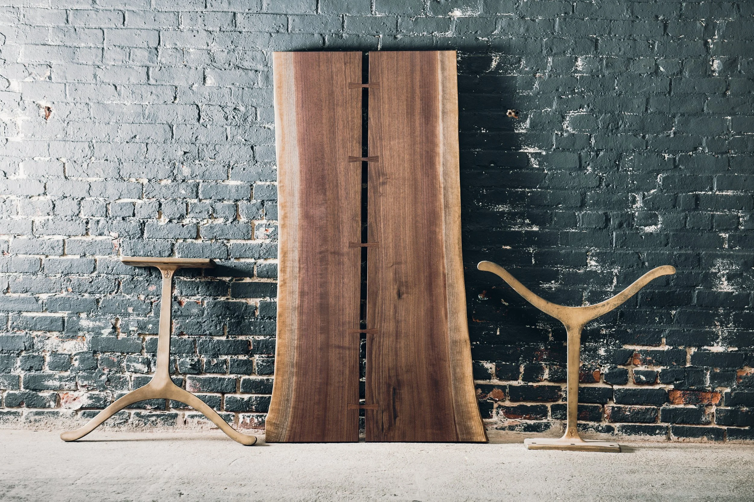 Two unfinished wooden tabletops and two wooden stands against a painted brick wall.