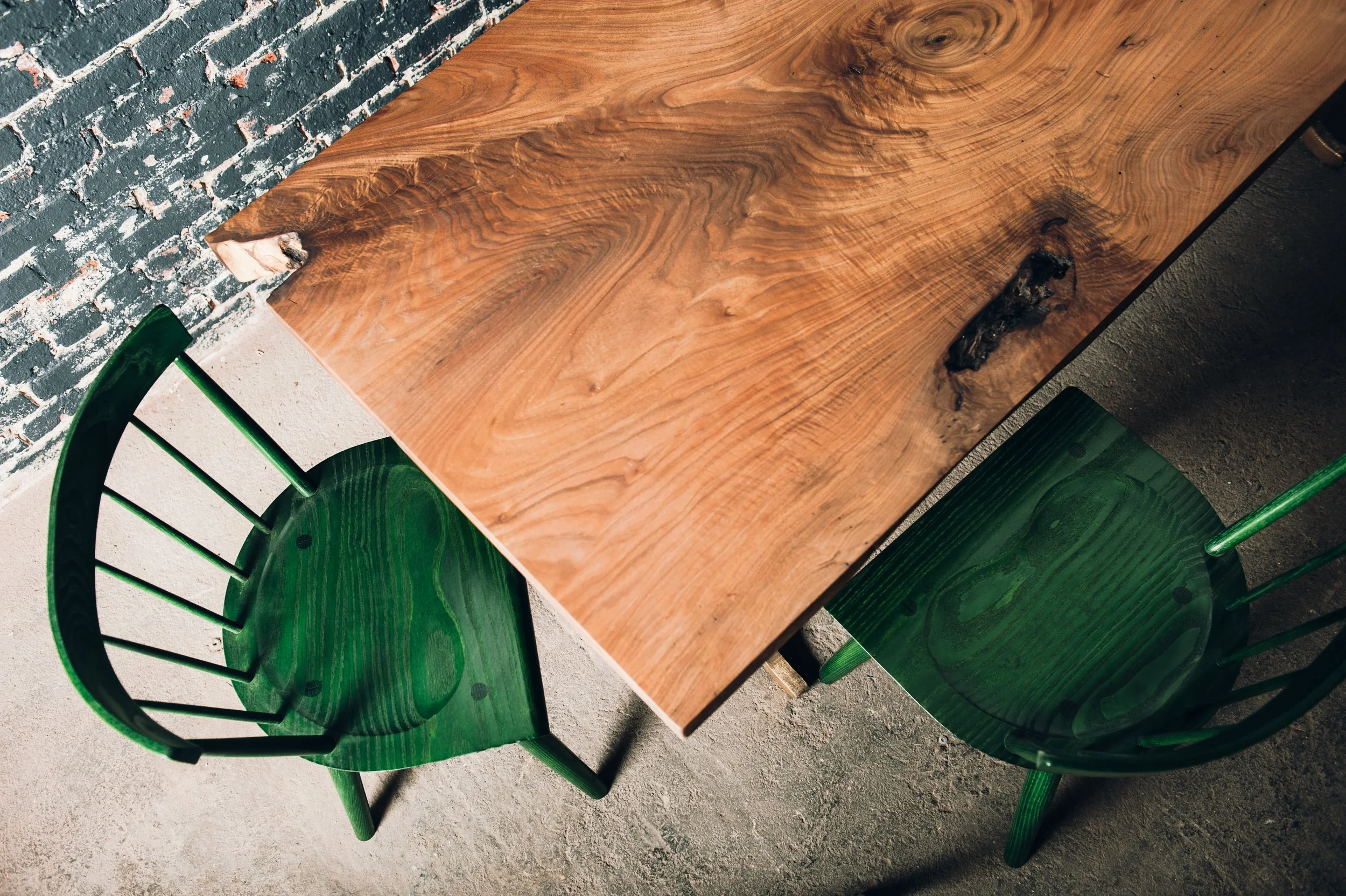 Top-down view of a wooden table with two green chairs placed on unpolished concrete floor, against a brick wall.