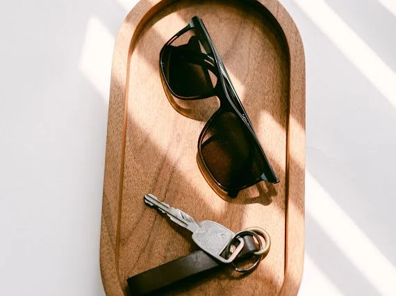Sunglasses and a set of keys with a keychain on a wooden tray with shadows from window blinds