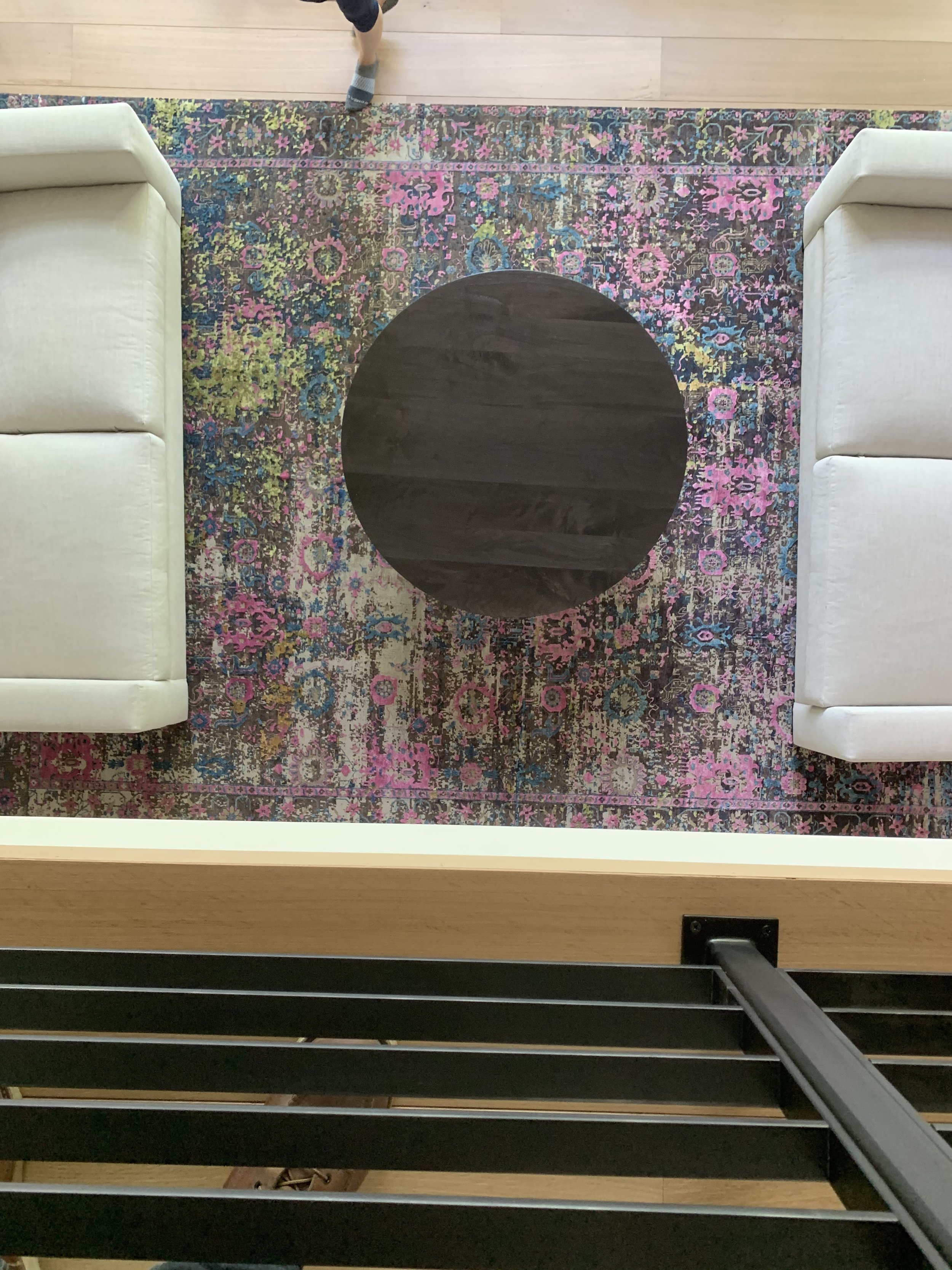 Top-down view of a living room area with a colorful pink, purple, blue, and green patterned rug, two white sofas, and a small black round table in the center. Part of a person's foot is visible at the top of the image.