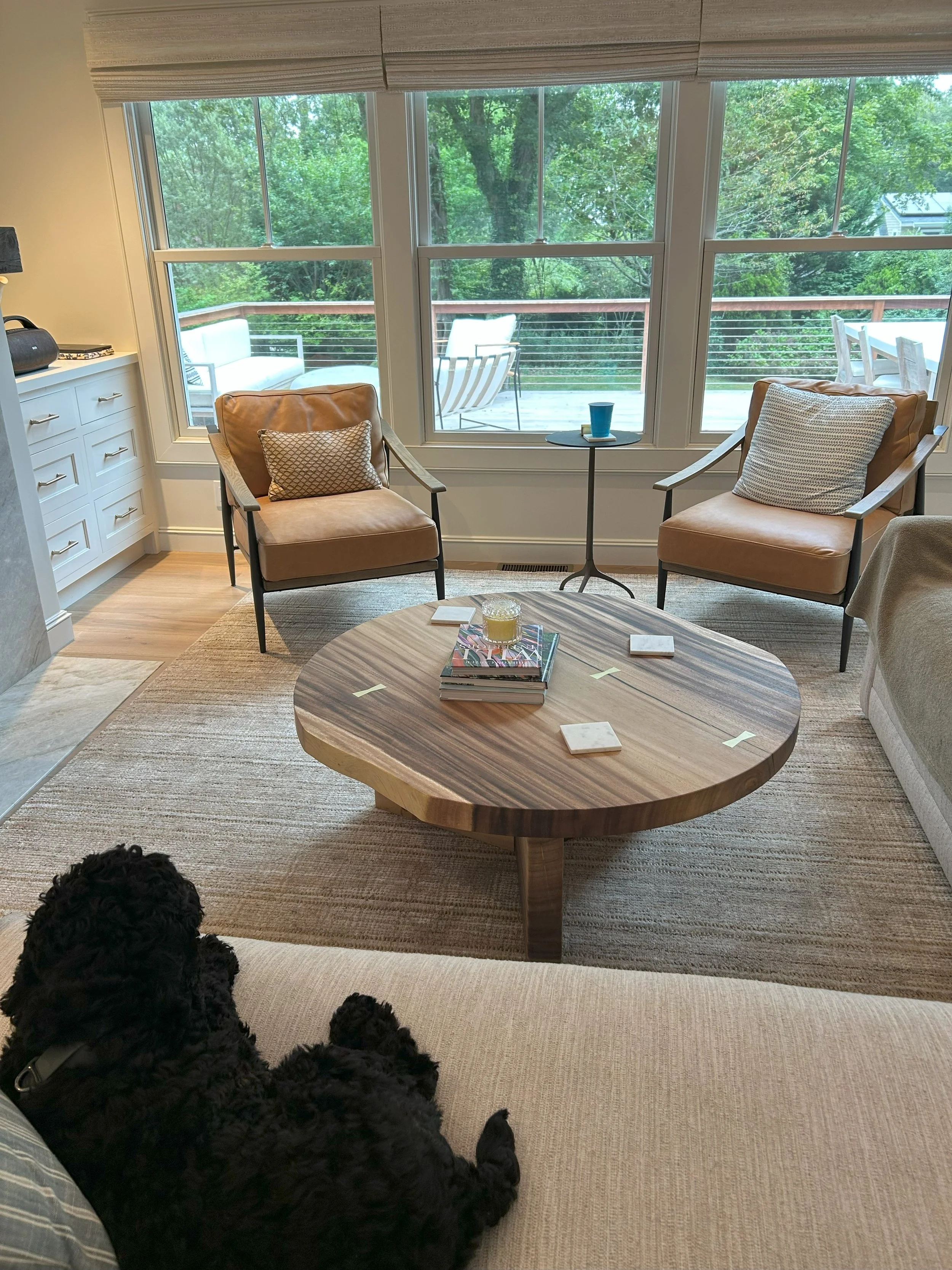 Living room with a large window overlooking a deck and trees outside. There are two tan chairs with pillows, a small round table with a blue cup, and a wooden coffee table with books and coasters. A black dog is lying on a beige sofa visible in the foreground.