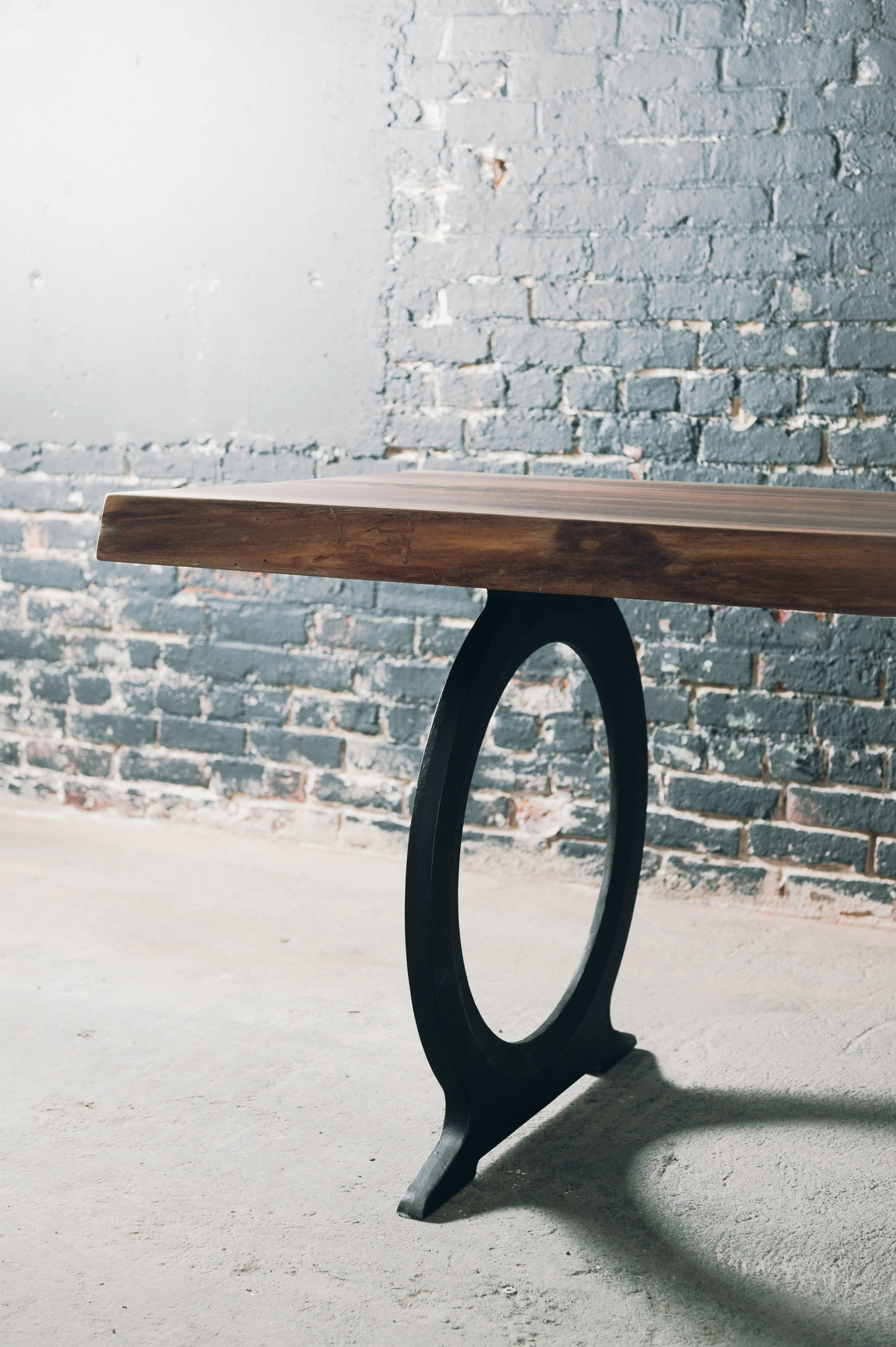 A modern wooden table with a circular black metal leg, set against a gray brick wall with a concrete floor.