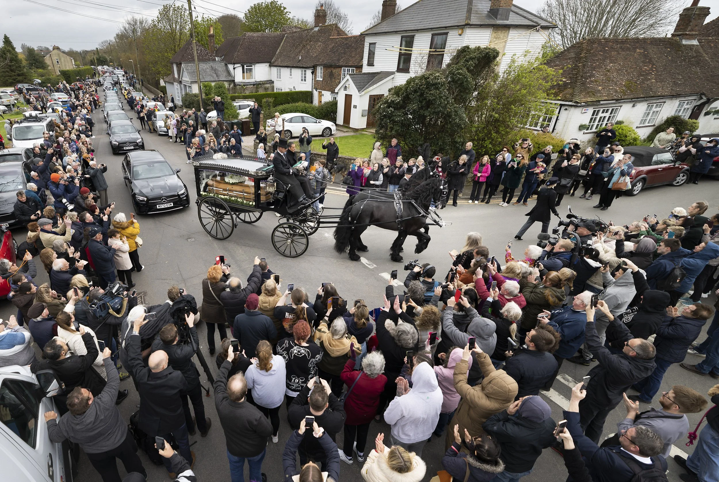 Crowds watch as a horse drawn hearse carrying the coffin of Paul O'Grady passes through the village of Aldington in Kent on April 20, 2023.