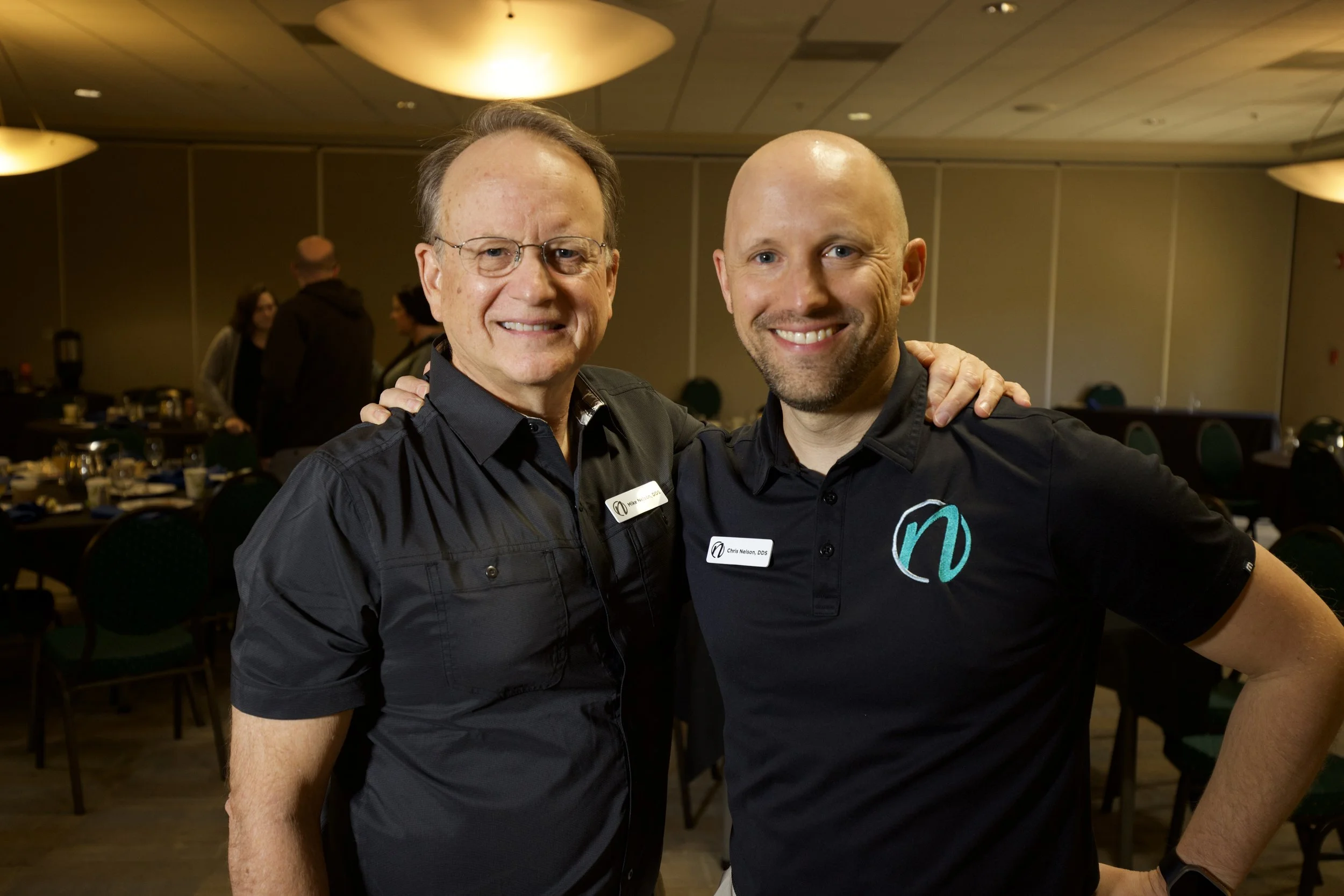 Two smiling men at a social event, with one man wearing glasses and a name tag that reads 'Chris Nelson, DDS', and the other man with a shaved head, both wearing black shirts, with a banquet room setting in the background.  Dr. Chris Nelson and Dr. M