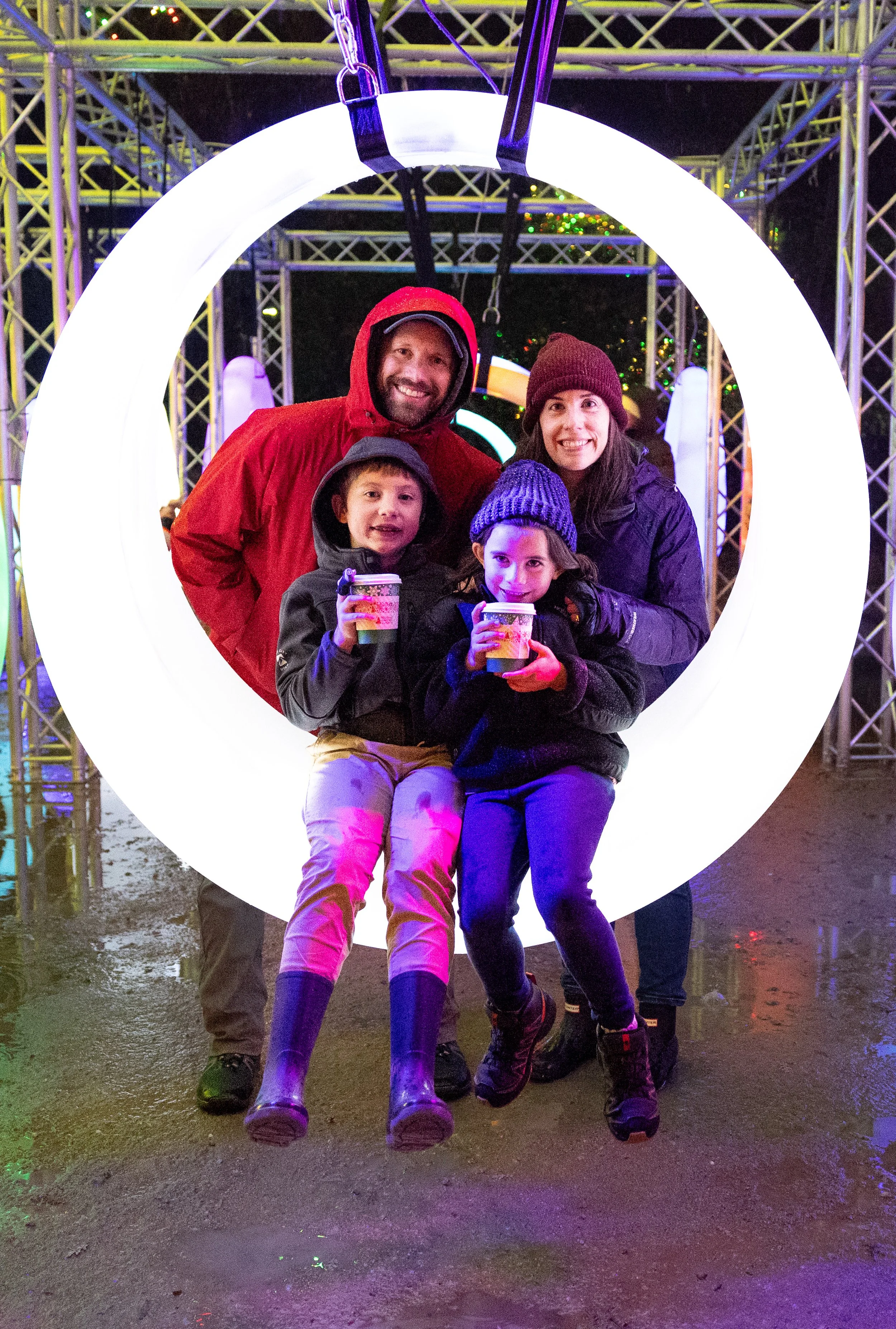 A family of four, dressed in warm clothes, standing inside a large glowing ring at night with illuminated metal scaffolding and green holiday lights in the background. The adults are smiling, and two children at Garden of Lights Redding