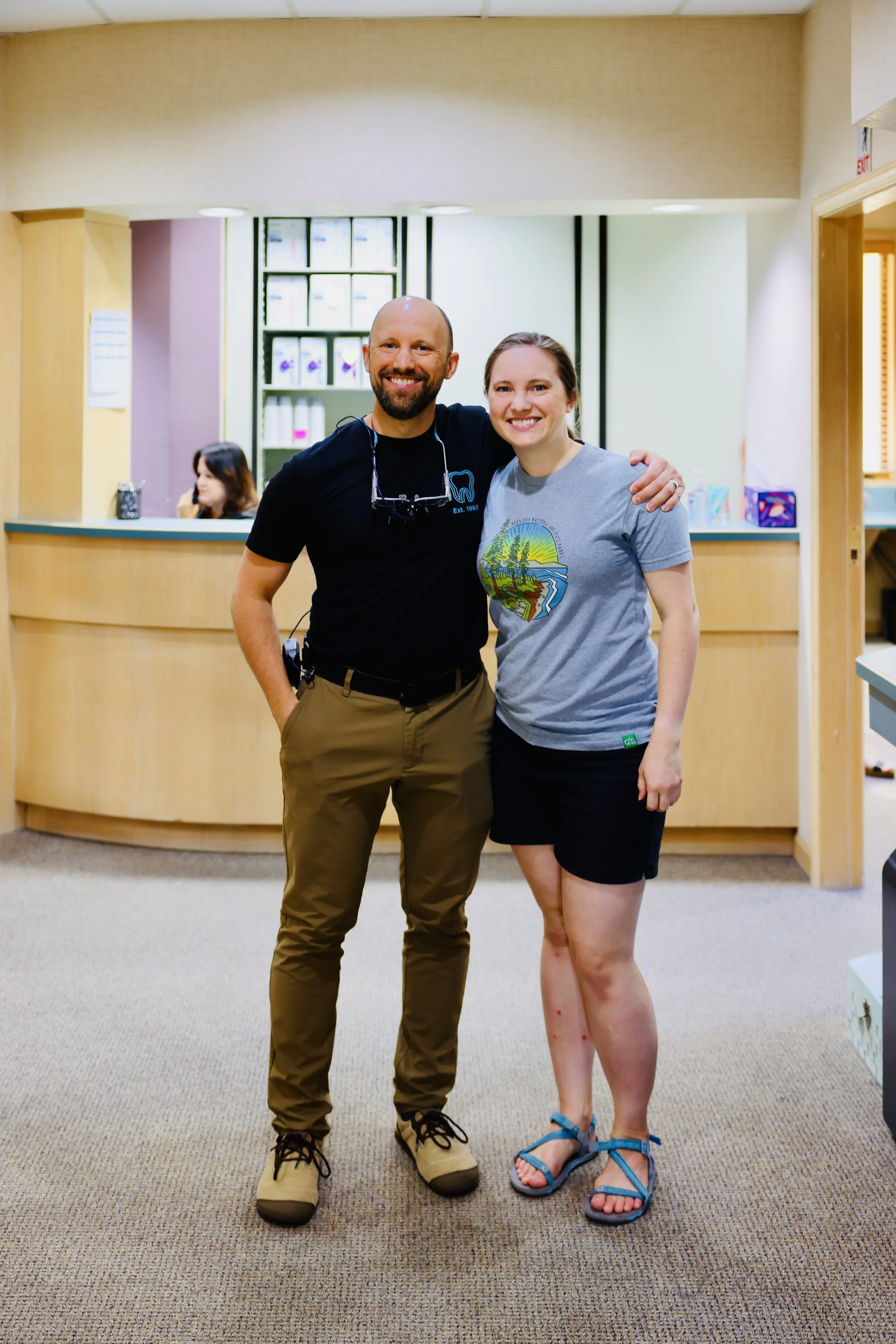 A man and woman standing together in a reception area, smiling at the camera. The man is wearing tan pants, a black T-shirt, and tan shoes, with a headset around his neck. The woman is wearing a gray T-shirt with a colorful design and black shorts, w