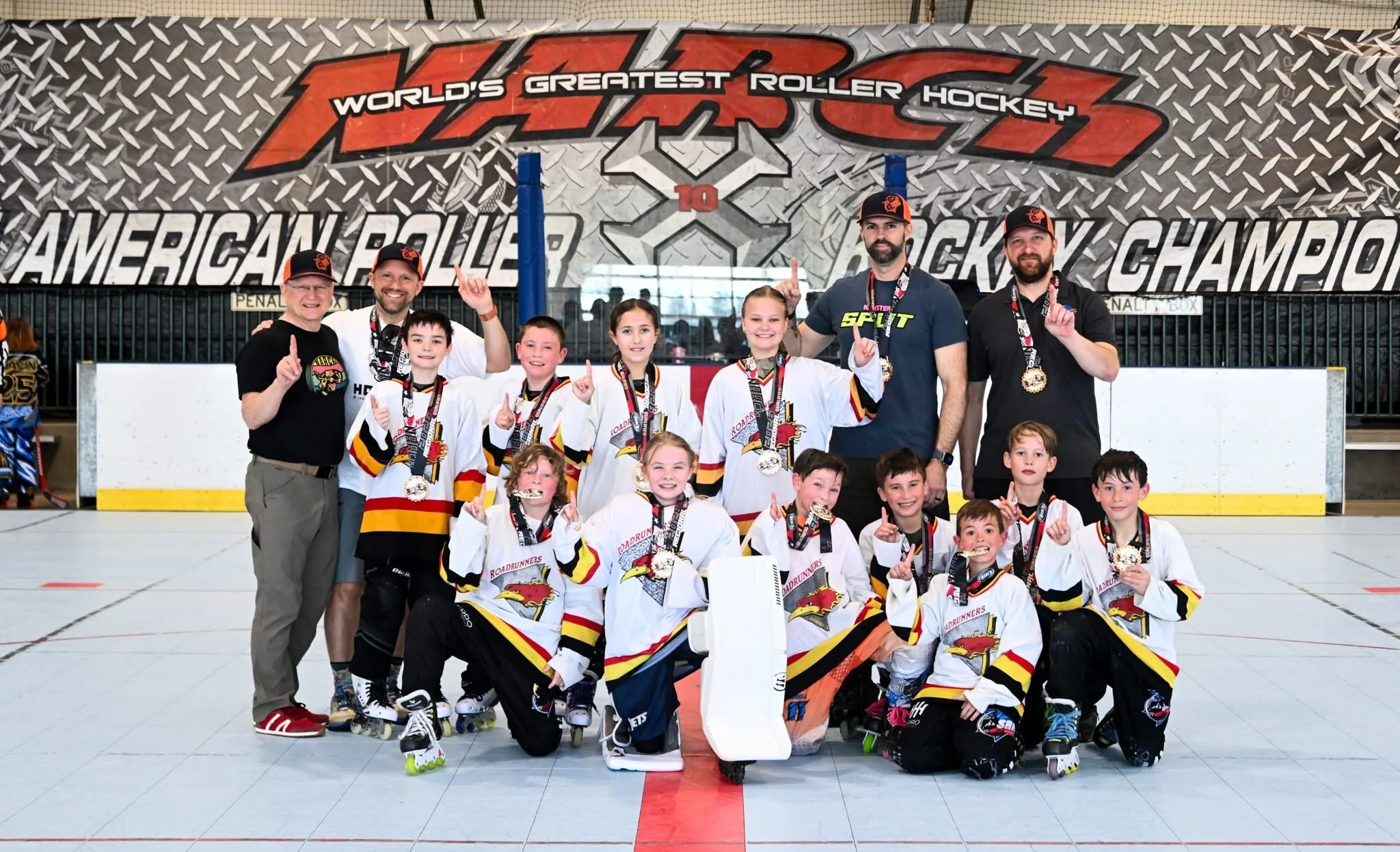 A hockey team with coaches and children posing with medals in an ice rink in front of a banner that reads "World's Greatest Roller Hockey" and "American Roller Hockey Championship." The team members are dressed in white hockey jerseys with a dragon logo and are holding up their index fingers in celebration.