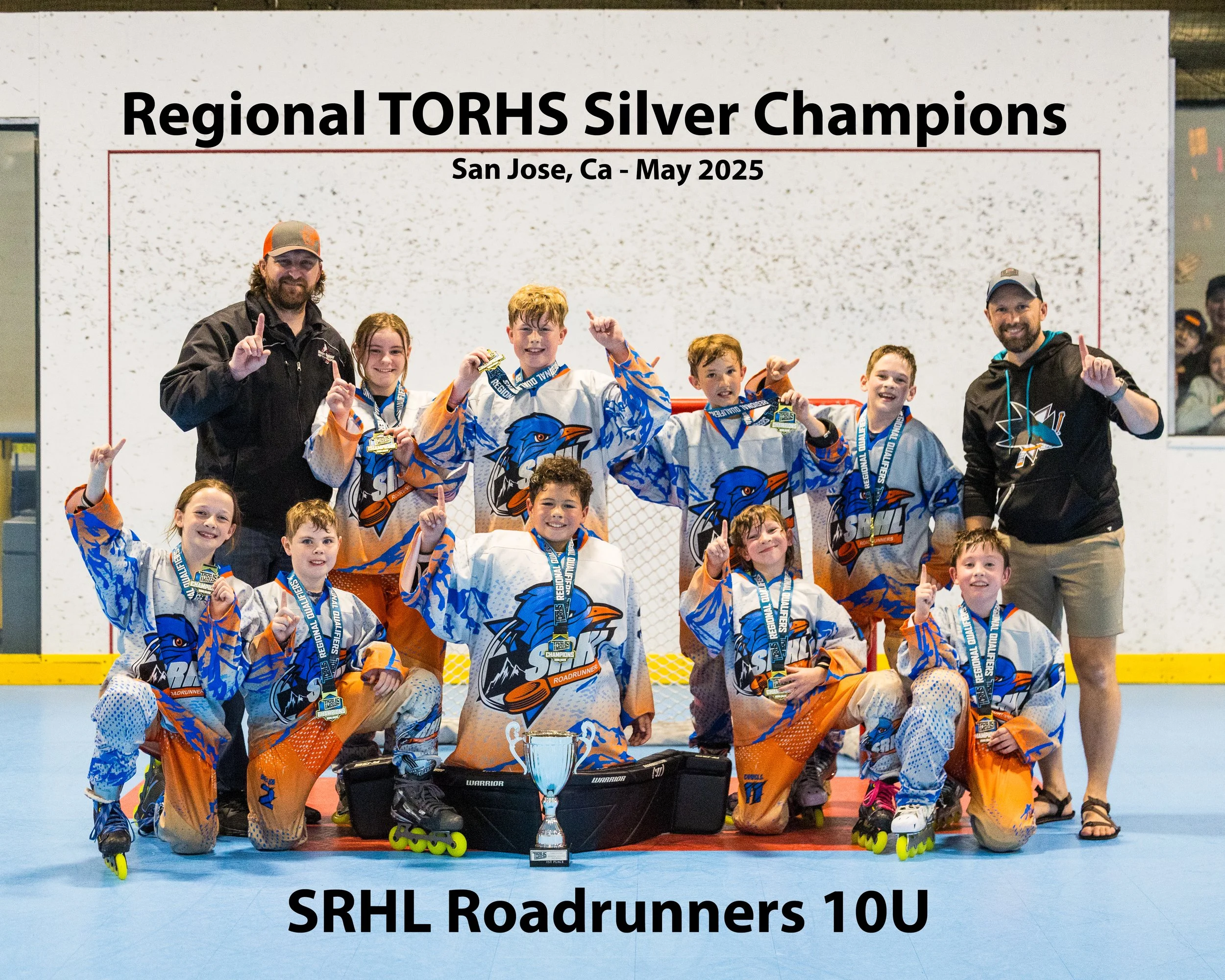 A group of young hockey players in white, blue, and orange jerseys celebrating with medals and a trophy at an ice rink, with two adults in black jackets and caps. A large sign behind them reads "Regional TORHS Silver Champions San Jose, Ca - May 2025